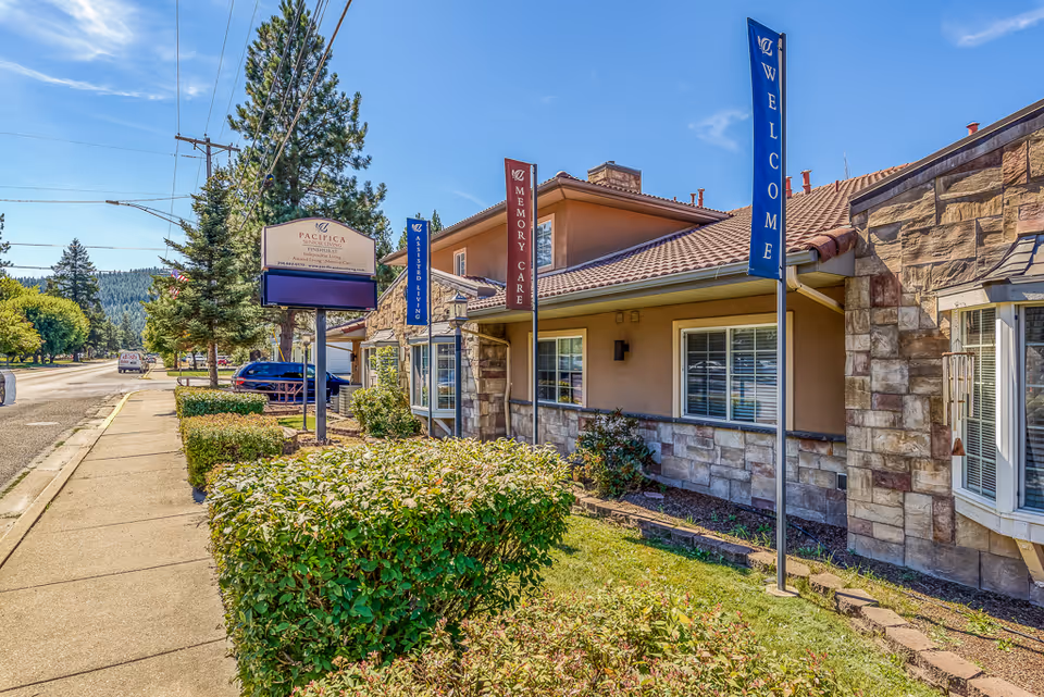 Exterior view of Pinehurst Senior Community building with stone and stucco facade, several flags reading 'WELCOME', 'MEMORY CARE', and 'ASSISTED LIVING', a sidewalk, bushes, and trees along the street under a clear blue sky.