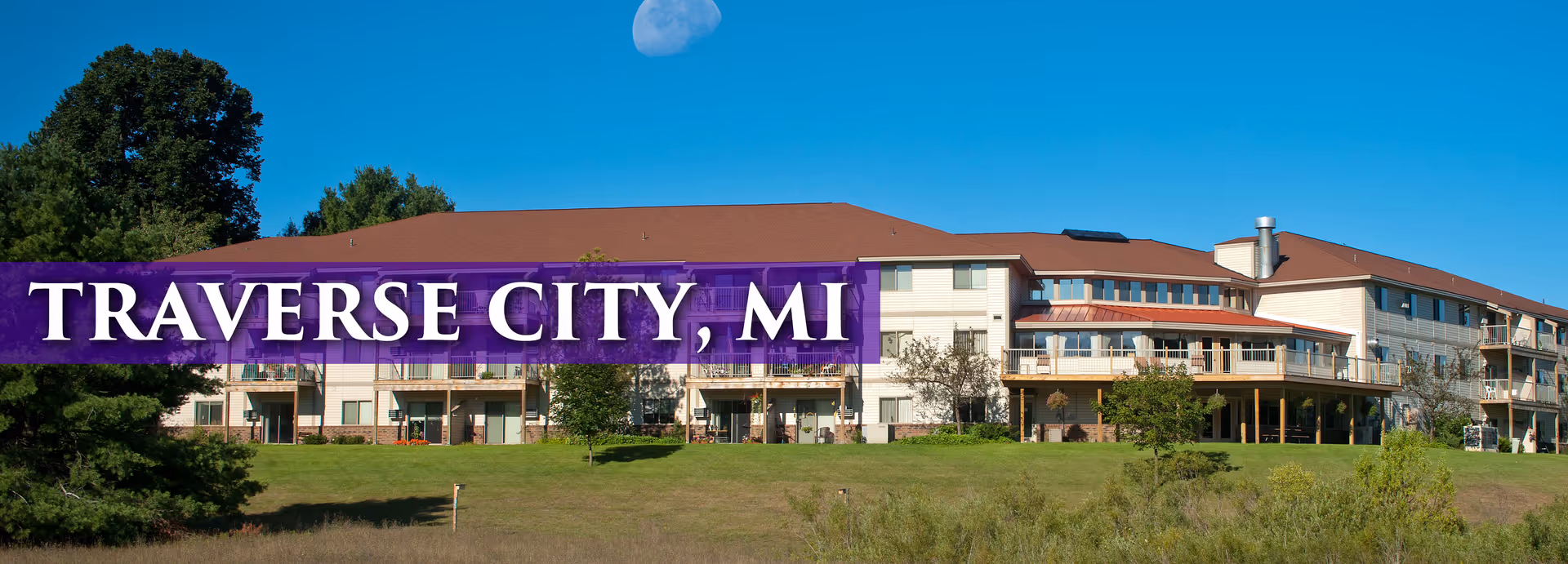 Three-story senior living building on a grassy lawn with a large purple banner reading "TRAVERSE CITY, MI" across the front.