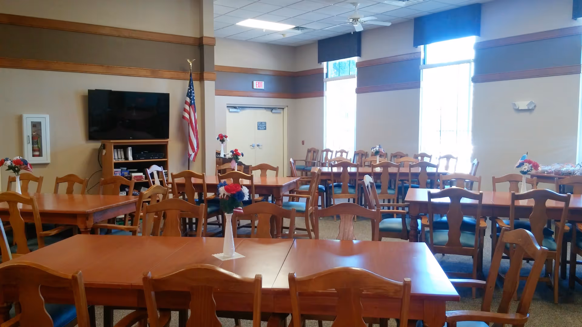 A dining room with multiple wooden tables and chairs arranged neatly. Each table has a small vase with red, white, and blue flowers and small American flags. There is a large flat-screen TV mounted on the wall next to an American flag. Large windows with blue valances allow natural light to brighten the room. An exit door is visible at the back.