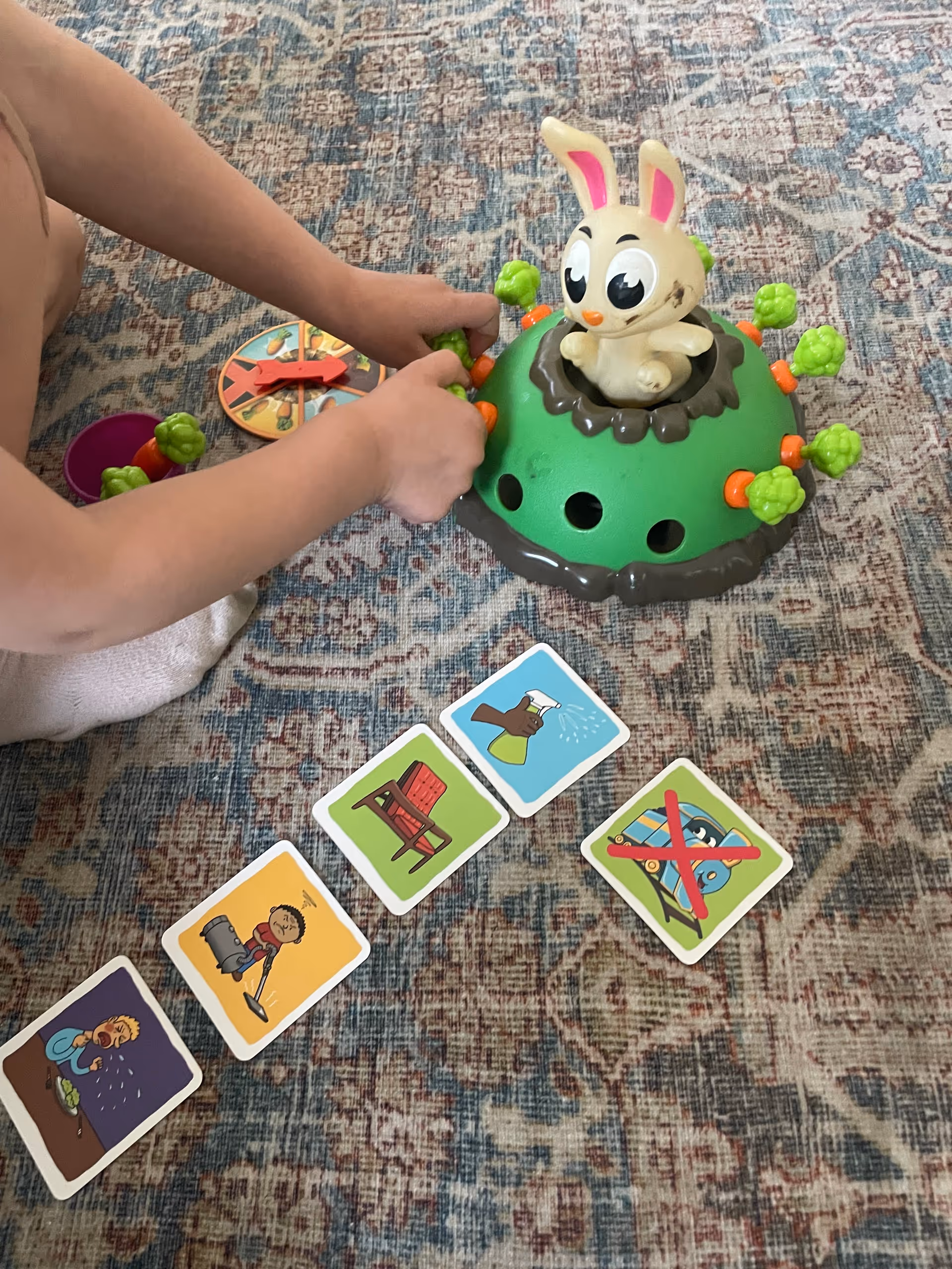 Child's hands operate a green bunny toy on a patterned rug with nearby cards.