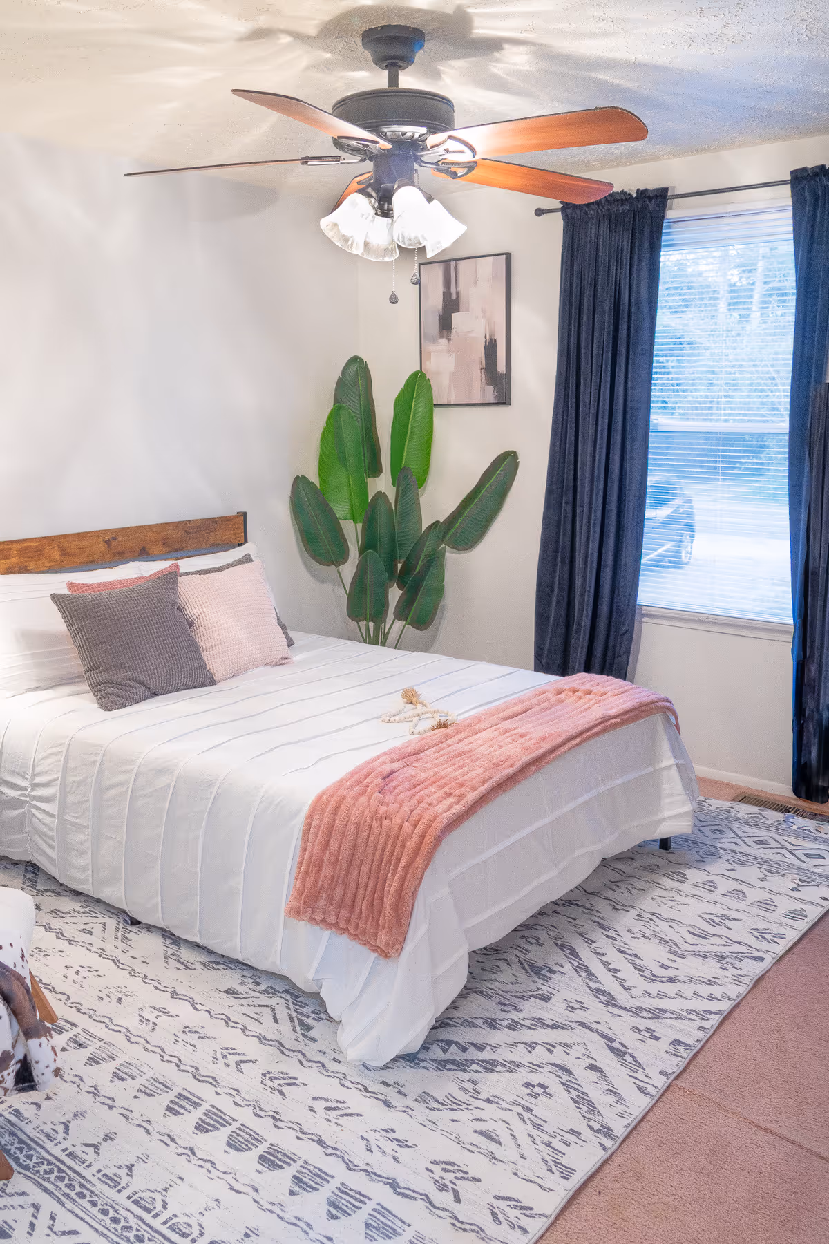 Bright bedroom with a bed, ceiling fan, plants, curtains, and a patterned rug.