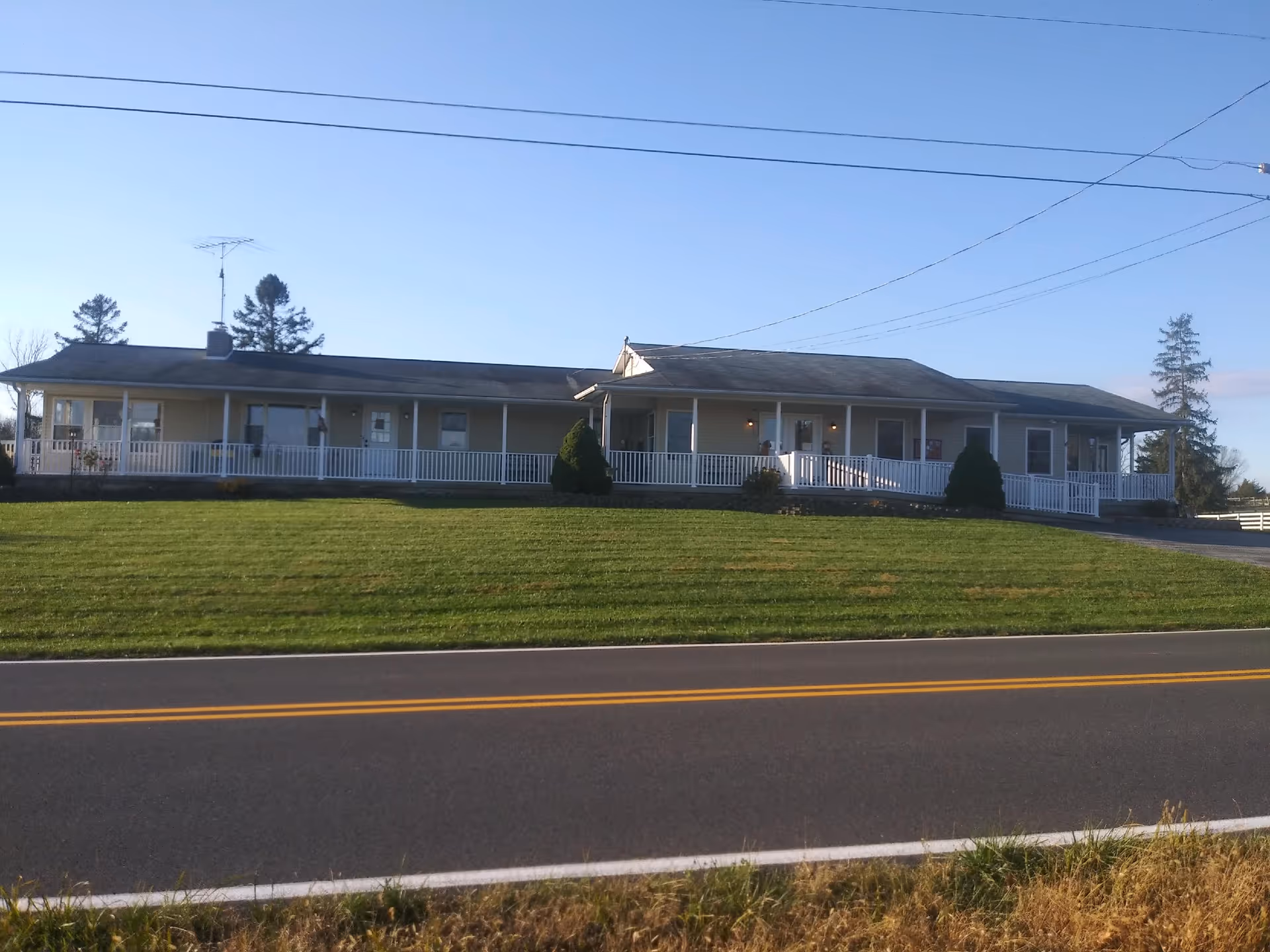 Long single-story building with a wraparound white railing porch, grassy lawn, and road in front.