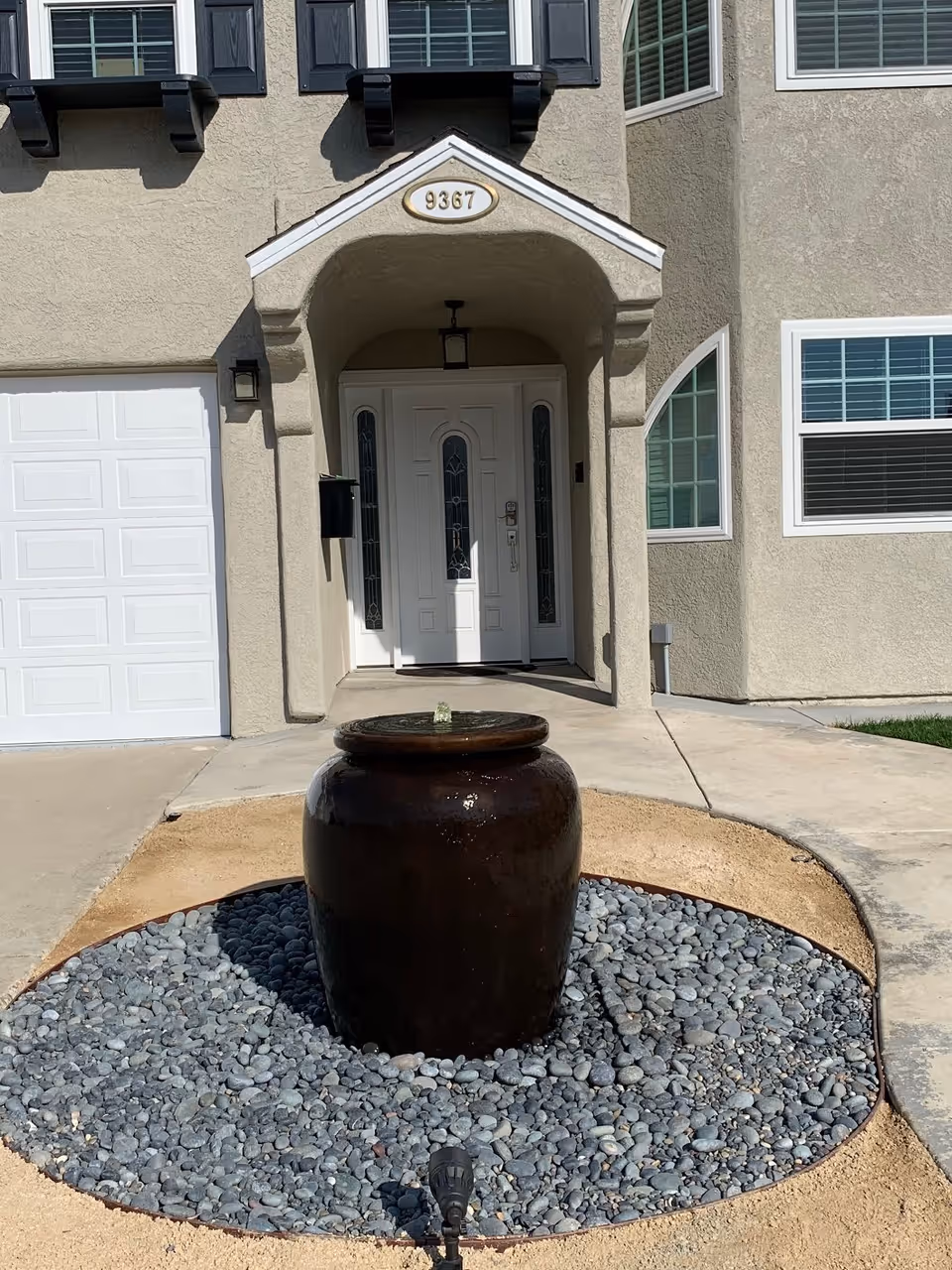 Front entrance of a building with a white door and decorative glass panels, a small covered porch with the number 9367 above it, and a large dark brown water fountain surrounded by gray pebbles in the foreground.