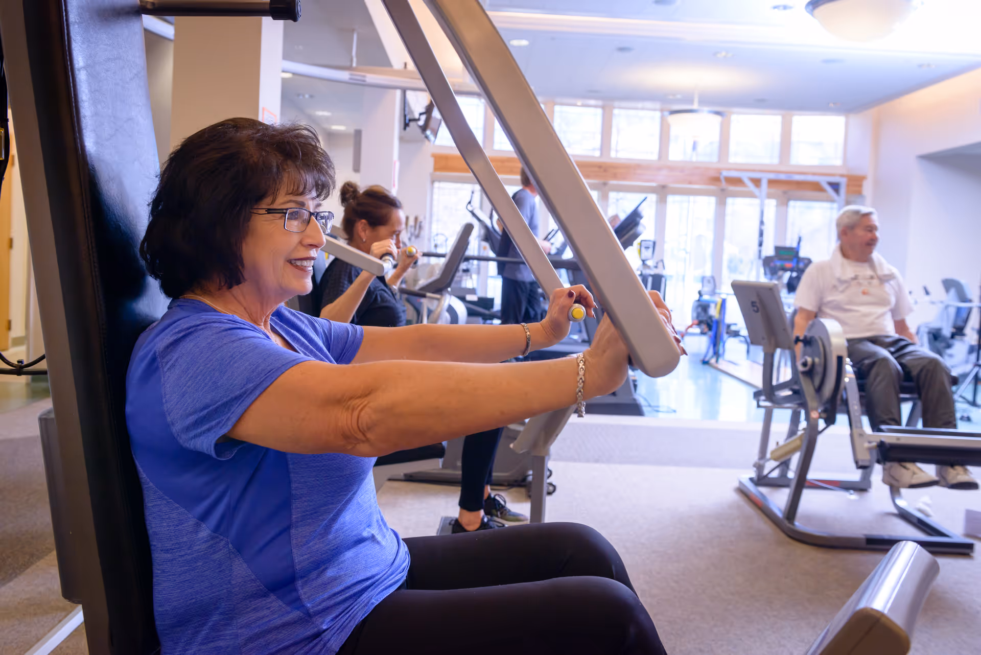 An elderly woman in a blue shirt using a chest press exercise machine in a fitness room. In the background, another woman is exercising with dumbbells and an elderly man is seated on a rowing machine. The room has large windows letting in natural light.