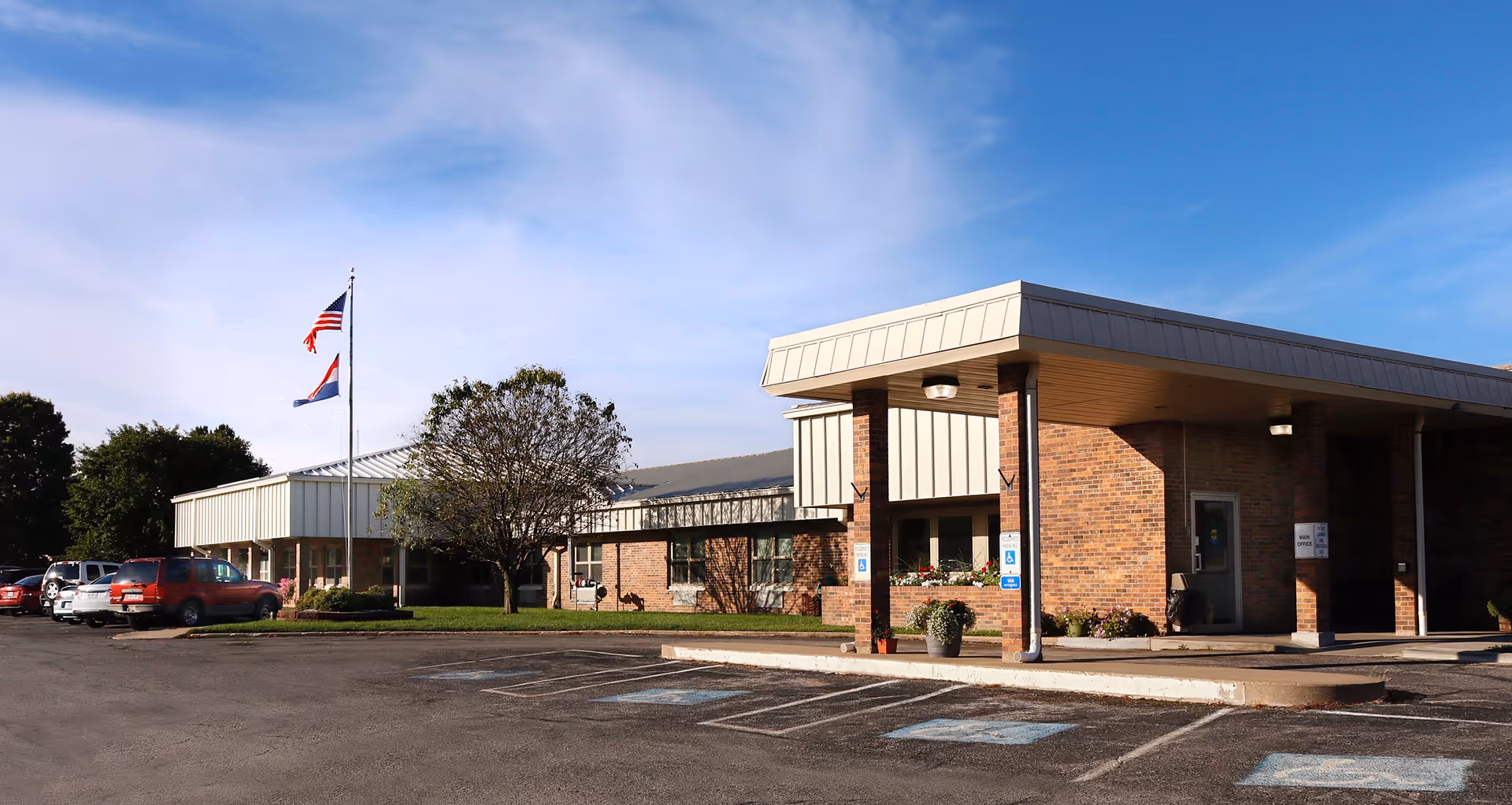 Exterior view of Shirkey Nursing & Rehabilitation Center showing a single-story brick building with a covered entrance, several parked cars, two flagpoles with American and state flags, and a clear blue sky.