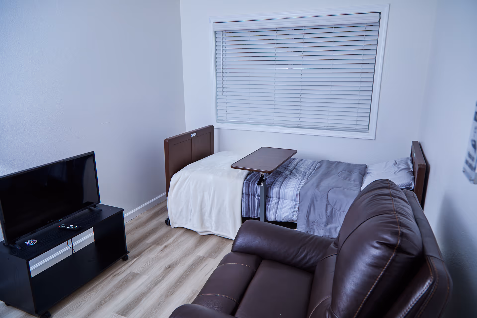Small furnished bedroom with bed, recliner, TV stand, and window blinds.