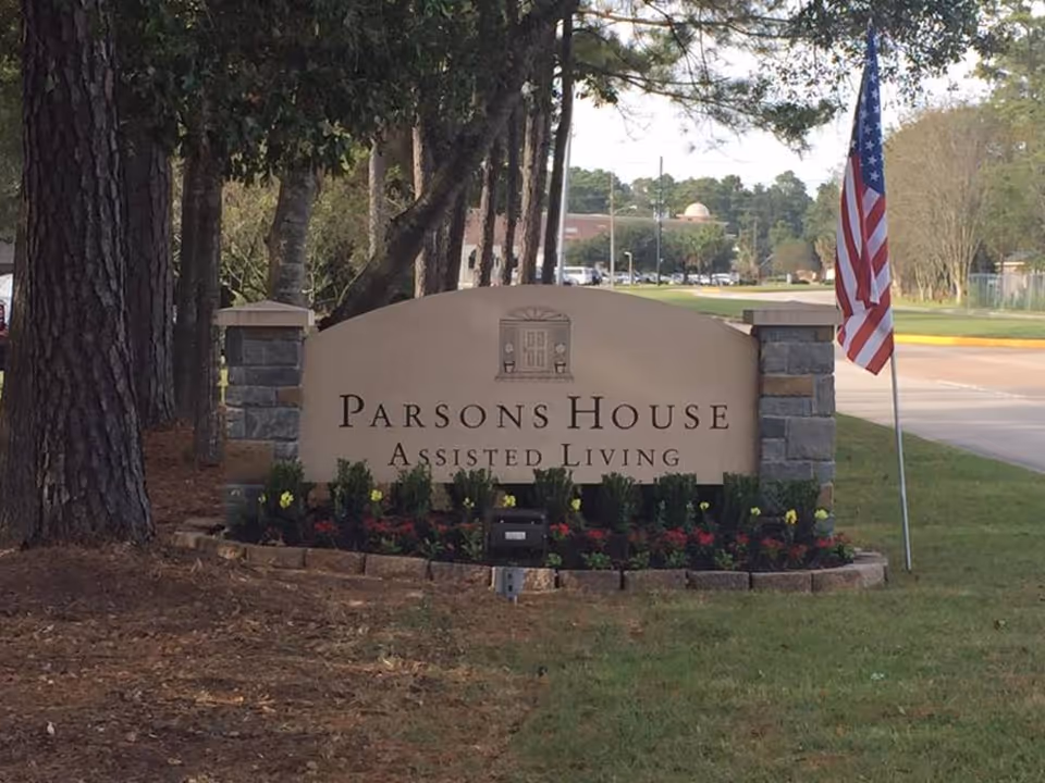 Stone and stucco sign for Parsons House Assisted Living surrounded by flowers and greenery, with an American flag on a pole to the right and trees in the background.