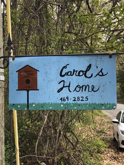 Blue sign reads Carol's Home with a birdhouse, outdoors near a road.