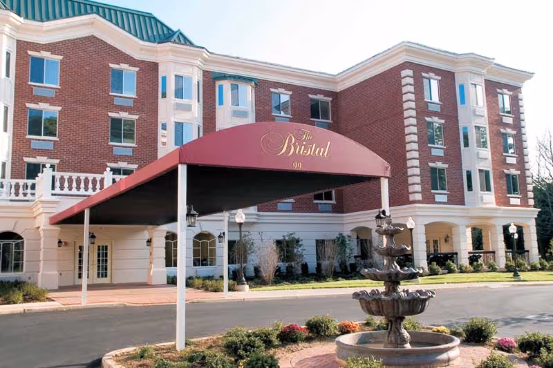 Front exterior of a red-brick assisted living building with a maroon entrance canopy reading "The Bristal" and a fountain in the circular drive.