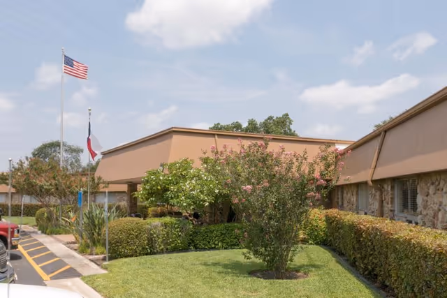 Exterior view of a single-story building with a tan roof and stone facade, surrounded by well-maintained landscaping including bushes, a flowering tree, and a green lawn. Two flagpoles display the American flag and another flag. The sky is partly cloudy.