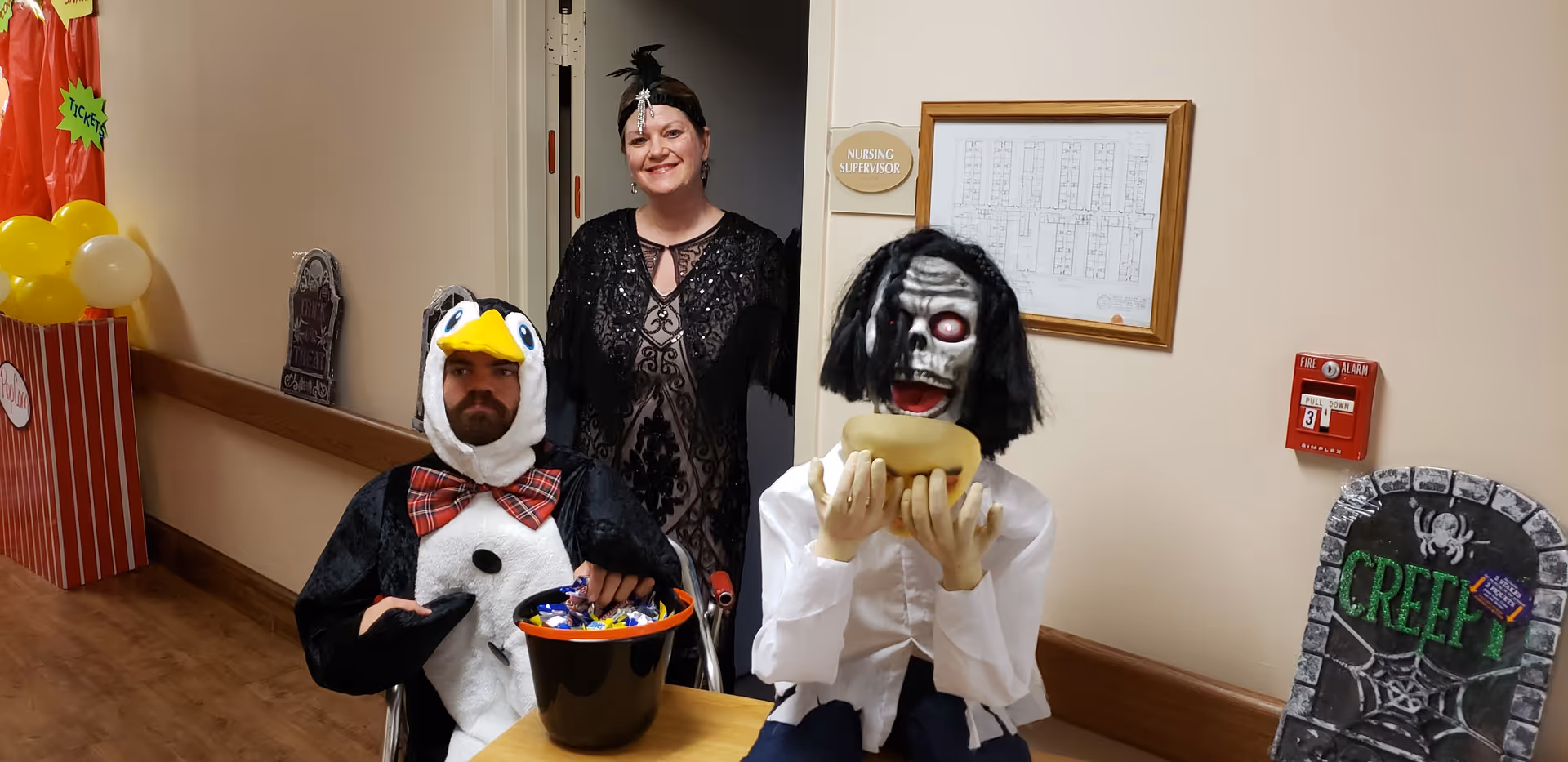 A man dressed in a penguin costume sitting in a wheelchair holding a bucket of candy, a woman standing behind him wearing a black dress and headpiece, and a Halloween decoration of a zombie-like figure holding a hot dog. The setting is a hallway with Halloween decorations including a tombstone with the word 'CREEPY' and balloons.