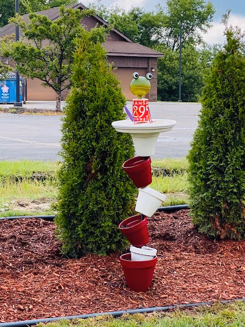 Colorful stacked buckets sculpture with a frog head sits among trees and mulch outside.