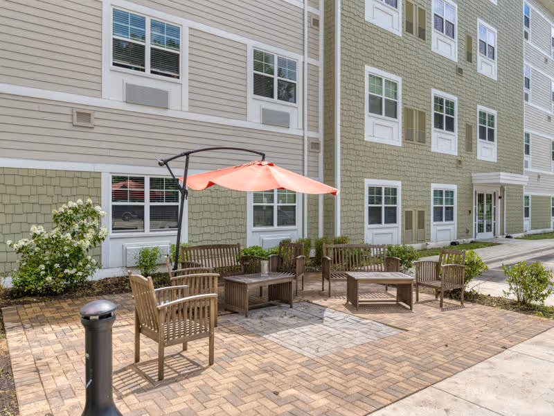 Outdoor patio area at a senior living facility with wooden chairs and tables arranged on a brick-paved surface. A large red umbrella provides shade over some of the seating. The building exterior is visible in the background with multiple windows and beige and green siding.
