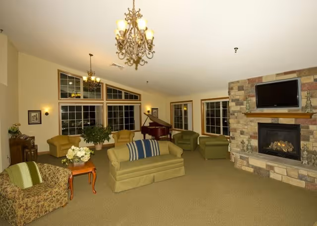 A spacious living room with beige carpet and cream-colored walls, featuring multiple armchairs and sofas arranged around a stone fireplace with a mounted flat-screen TV above it. There is a grand piano near large windows with multiple panes, and two ornate chandeliers hanging from the ceiling provide warm lighting. A small wooden side table with a flower arrangement is also visible.