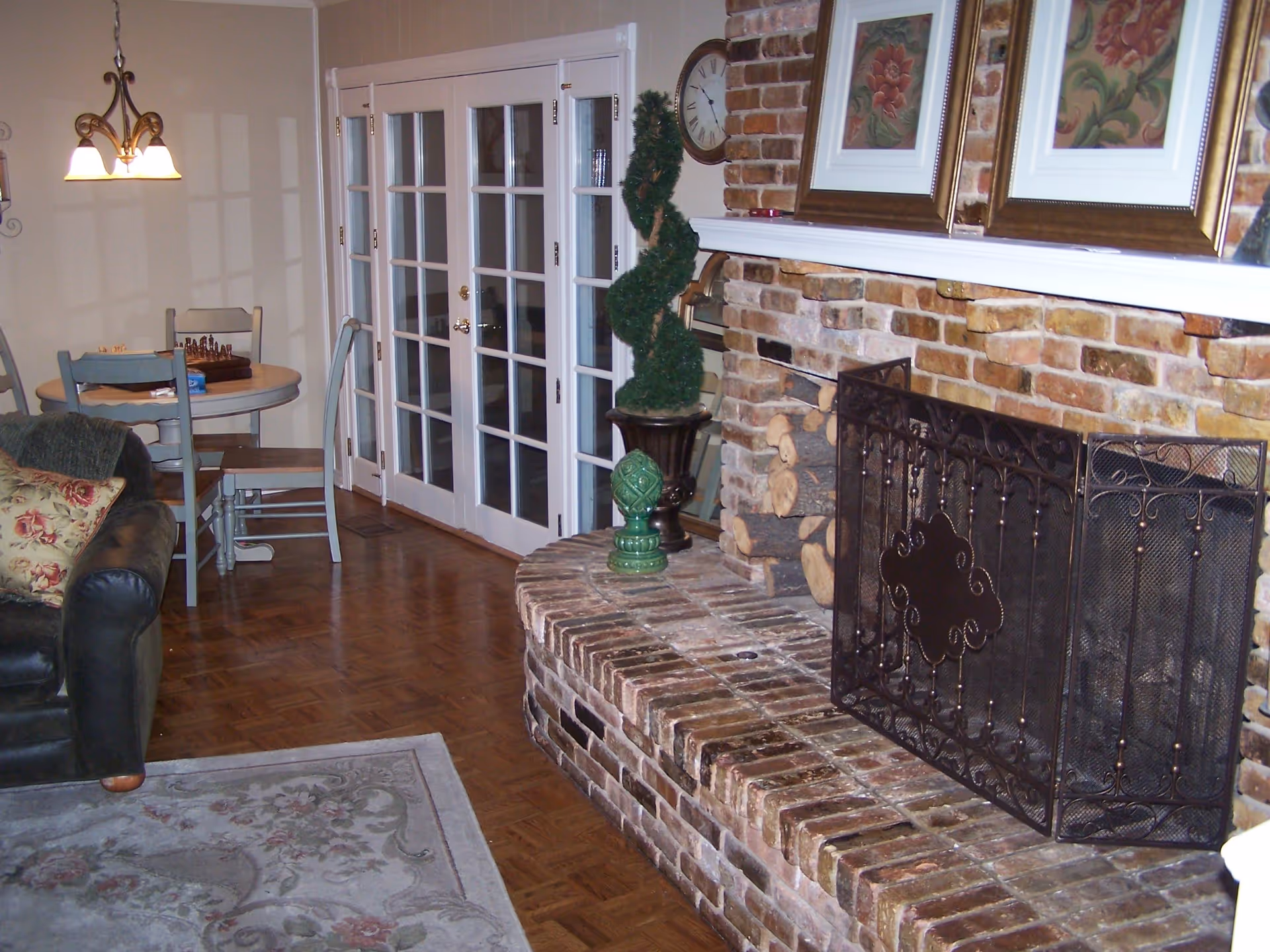 Cozy living room showing a brick fireplace with a decorative metal screen, a leather sofa, area rug, and a small dining table by French doors.