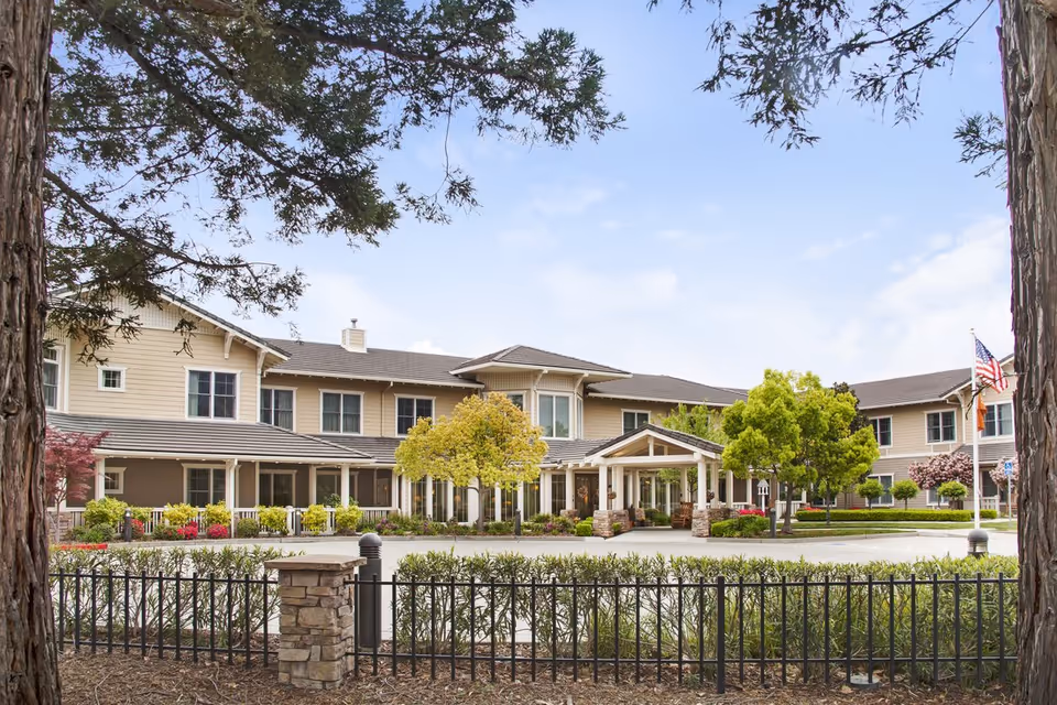 Front exterior of a two-story beige senior living facility with a covered main entrance, landscaped grounds, trees, and flagpoles.