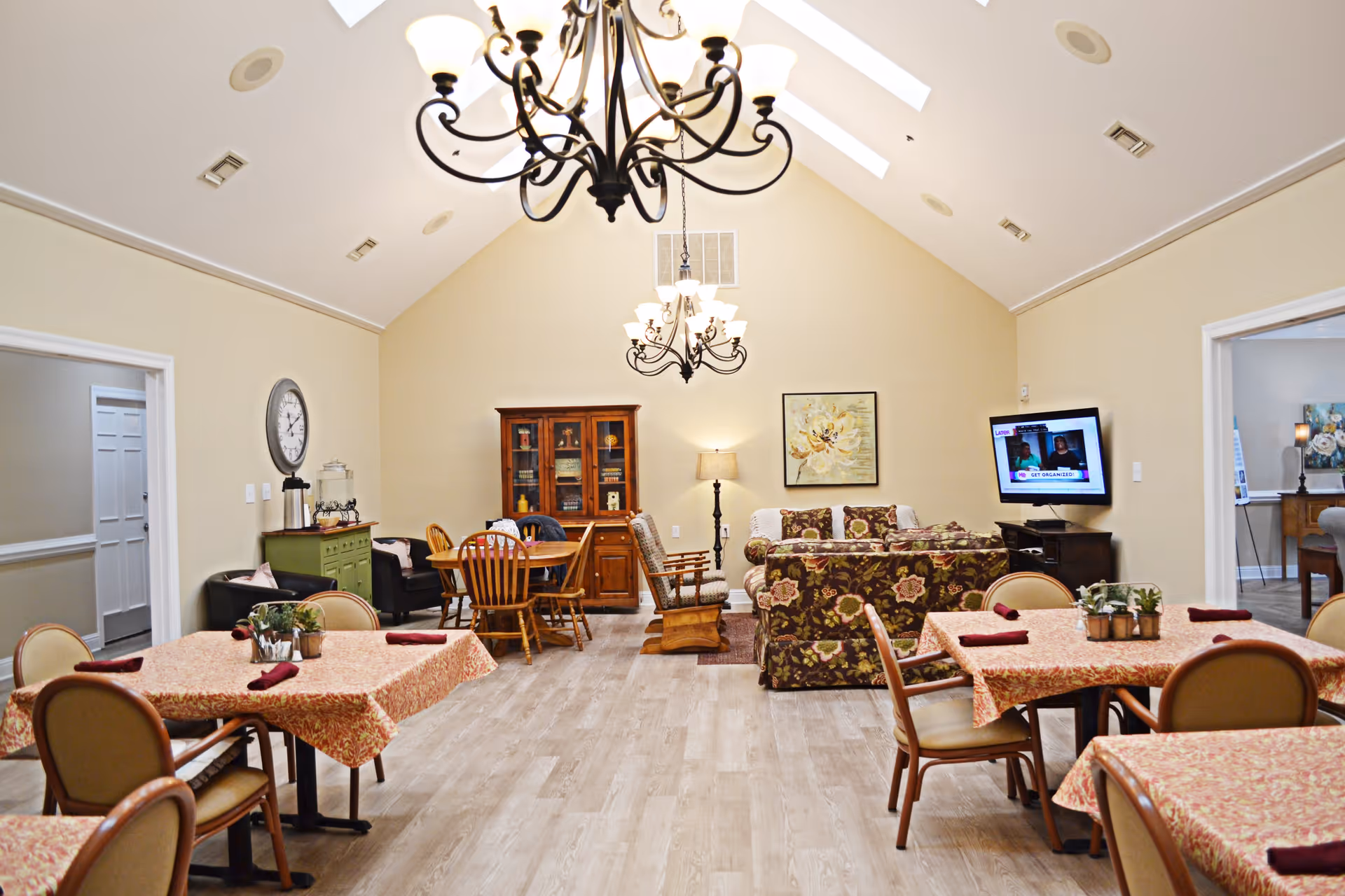 A bright and spacious common area with high vaulted ceilings and two chandeliers. The room features several tables with floral tablecloths and chairs arranged for dining, a sitting area with floral-patterned couches and a rocking chair, a wooden cabinet, a wall-mounted TV, and decorative wall art. The flooring is light wood, and there are doorways leading to other rooms.