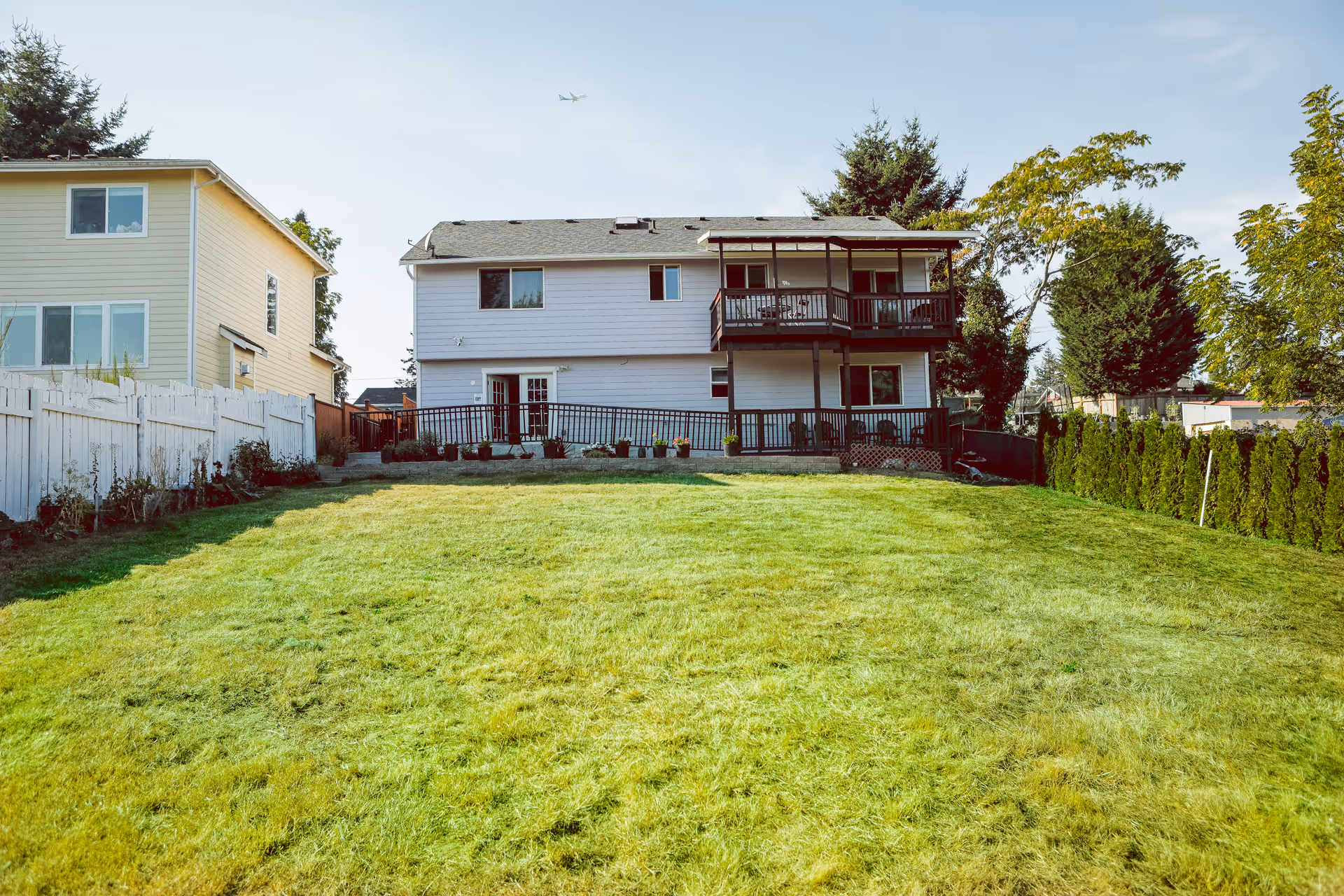 View of the backyard of a two-story house with a green lawn, a wooden deck with railings, and surrounding trees and bushes under a clear sky with an airplane flying overhead.