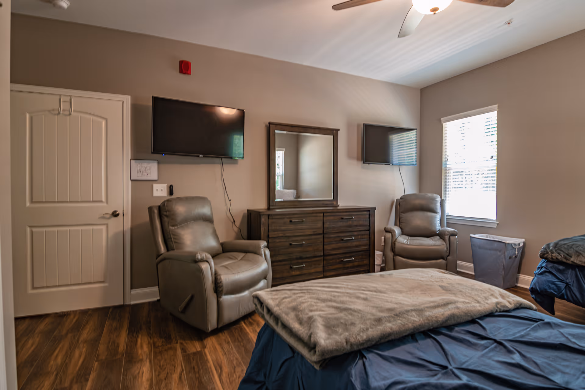 A furnished bedroom with two recliners, dresser with mirror, and wall-mounted TVs in a cozy suite.