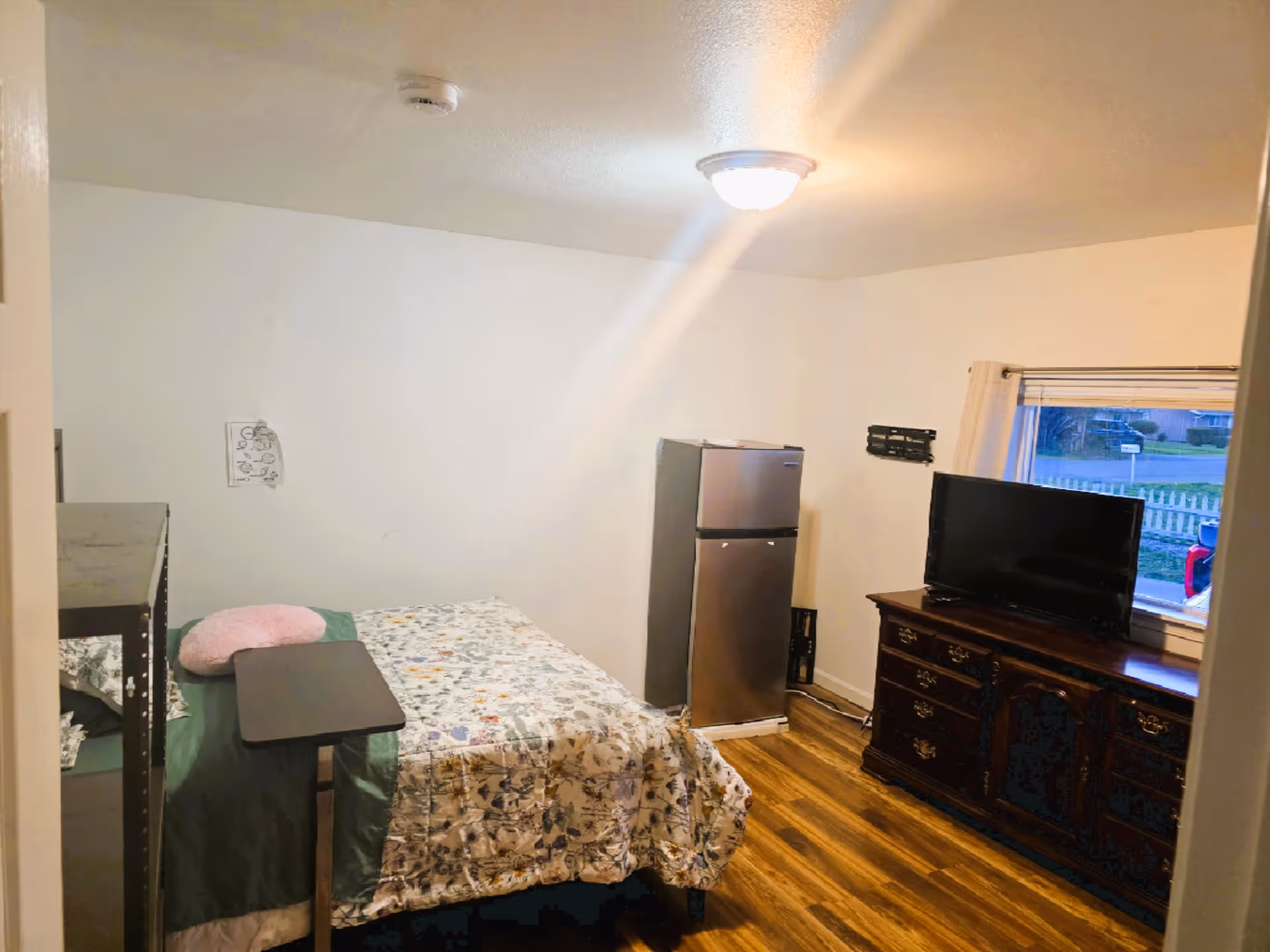Small bedroom with bed, dresser, TV, and a fridge by a window.