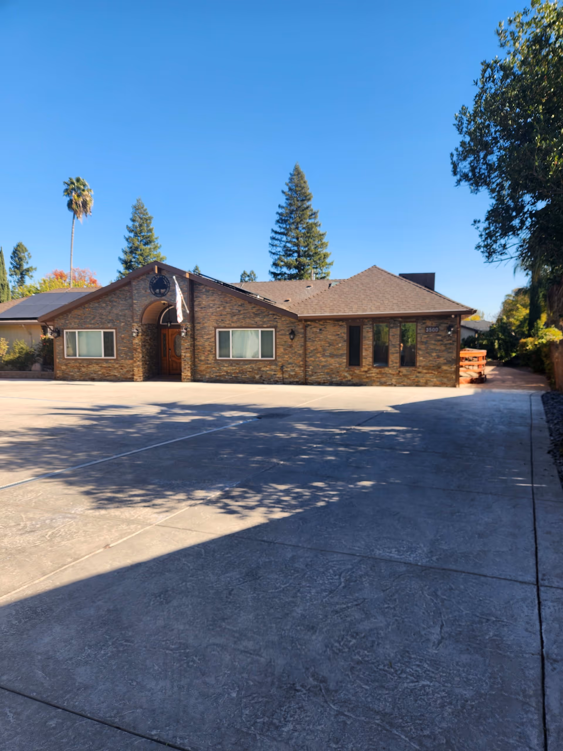 Exterior view of a single-story brick building with a sloped roof under a clear blue sky. The building has several windows and a wooden front door with an archway. There is a large concrete driveway in front and tall trees in the background.