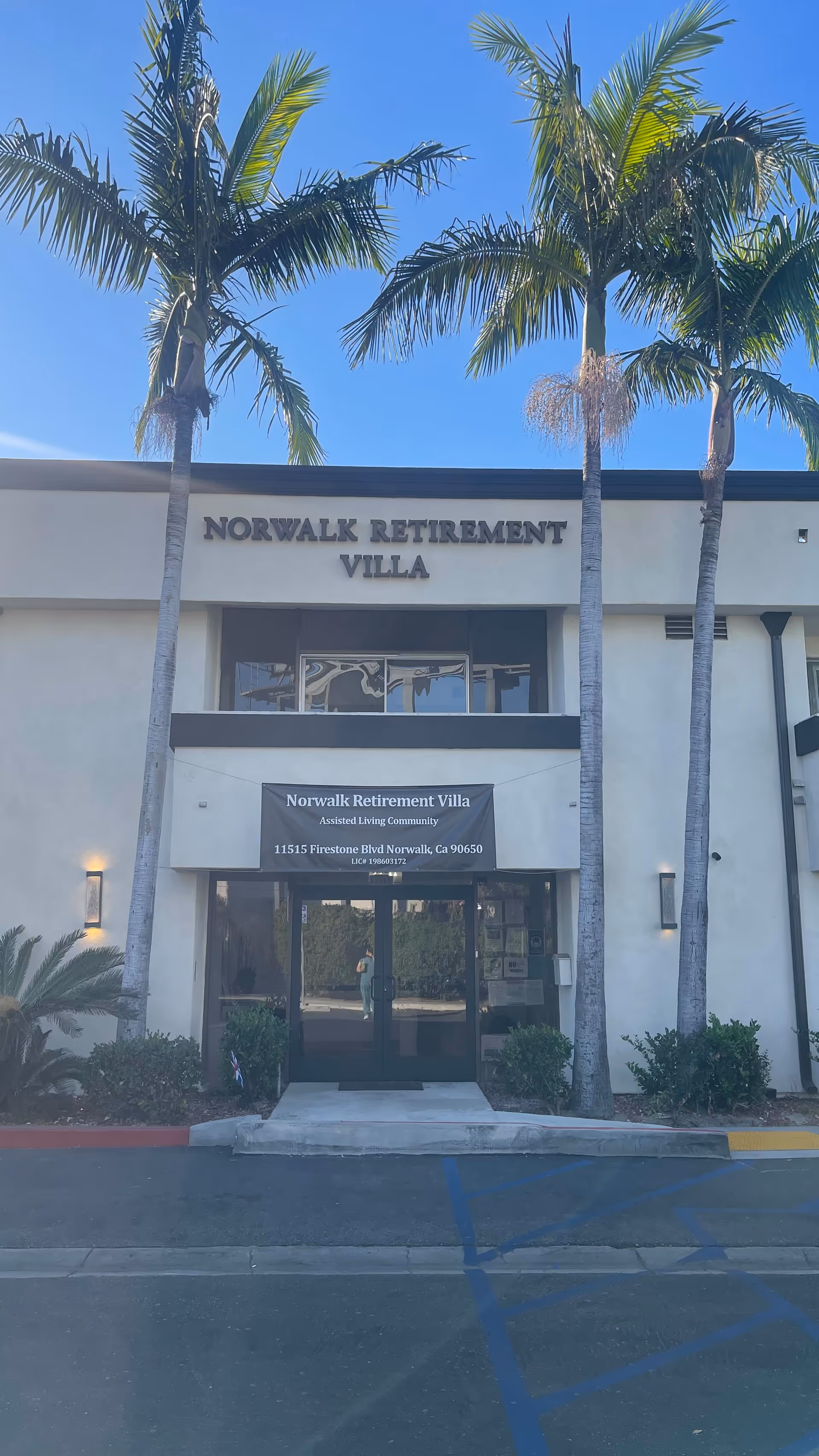Exterior front entrance of the Norwalk Retirement Villa building with palm trees and the facility sign above the doors.