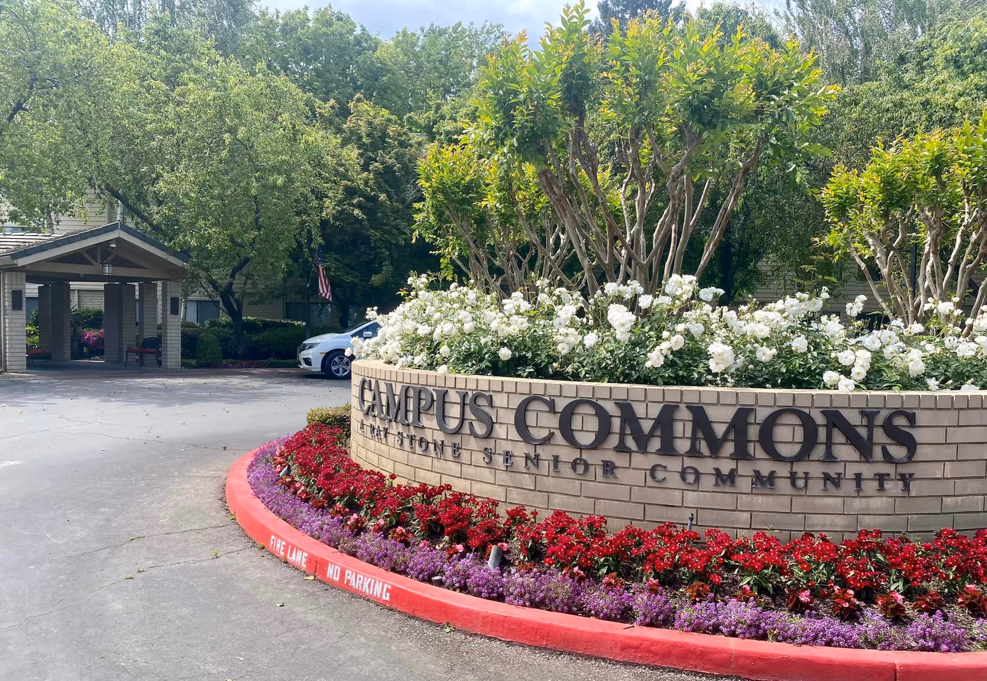 Entrance to Campus Commons senior living community with a brick sign surrounded by white and red flowers and green trees in the background. A covered driveway and a parked car are visible behind the sign.