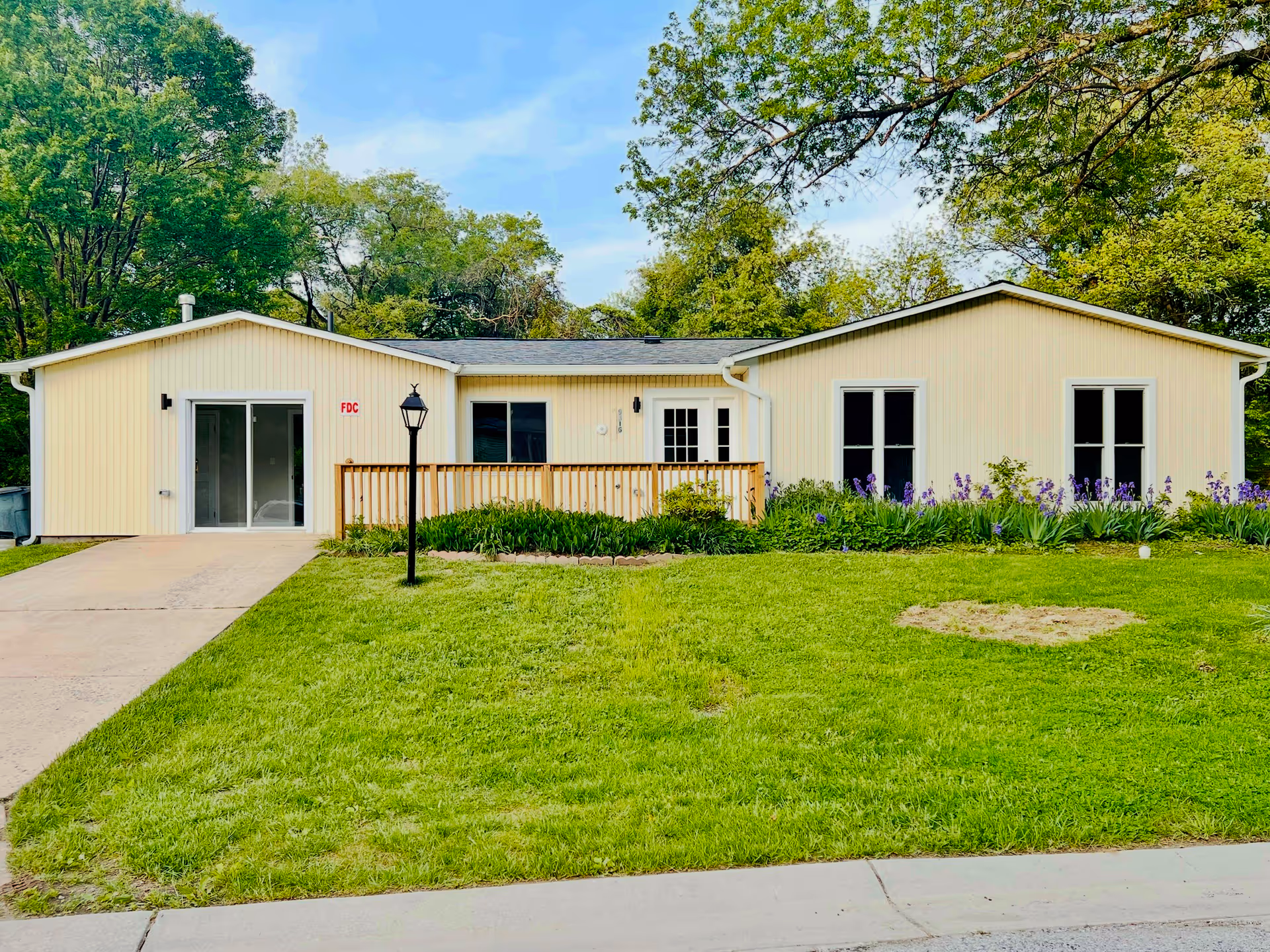 Single story yellow senior care home with green lawn, flower beds, and a wooden front deck.