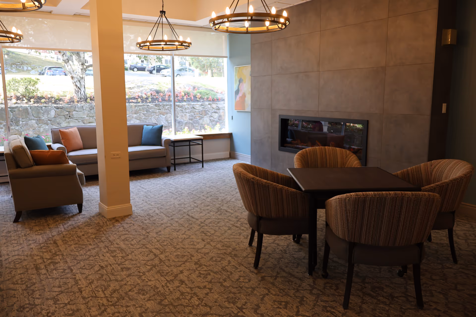 A cozy living room area with two gray sofas adorned with colorful throw pillows near large windows showing an outdoor view. There is a modern fireplace set into a tiled wall and a square table surrounded by four upholstered chairs with striped fabric. Two circular chandeliers hang from the ceiling, and the floor is covered with patterned carpet.