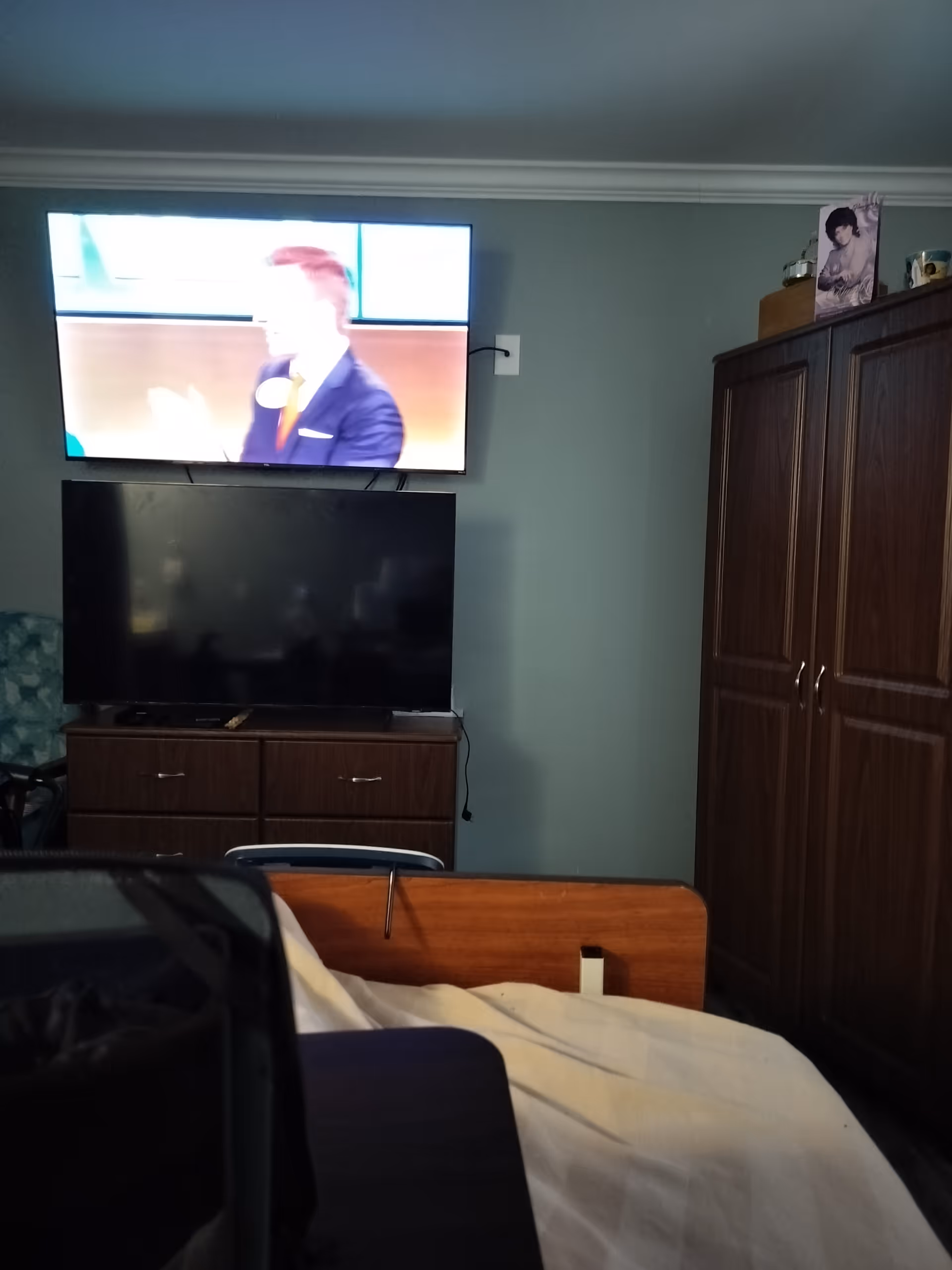 Senior living bedroom with a bed, dresser, large armoire, and TVs.