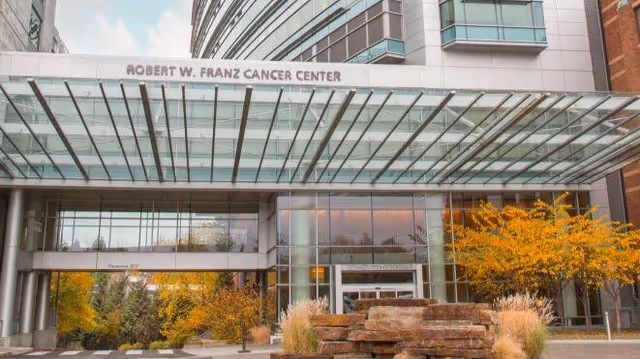 Hospital entrance with glass canopy, landscaping, and rock feature in front.