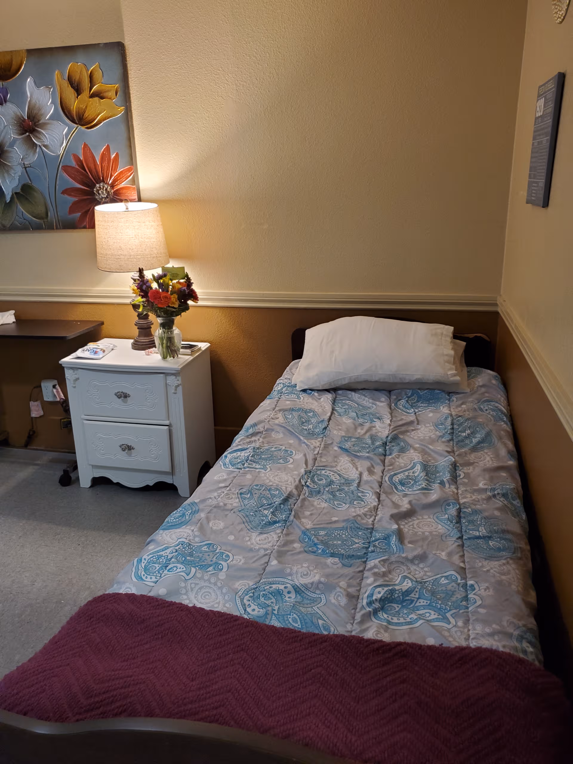 A single bedroom in a senior living facility featuring a bed, nightstand, lamp, and flowers.