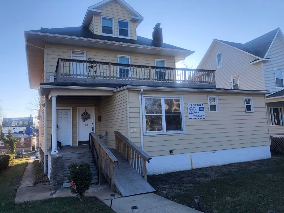 Two-story beige boarding house with ramp, front porch, and open-house sign.