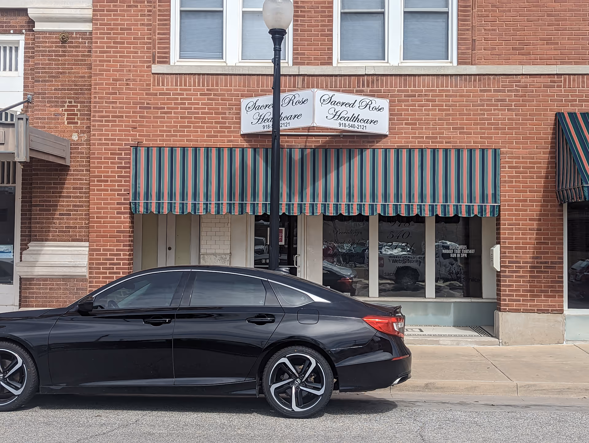 Brick storefront with striped awning, Sacred Rose Healthcare signage, and a black car parked outside.