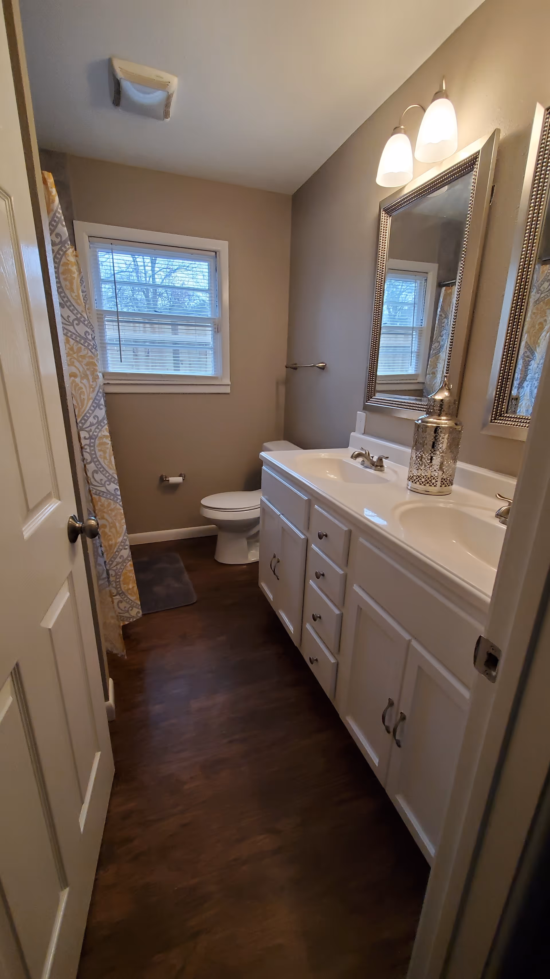 Bright bathroom with double vanity, framed mirrors, toilet, window, and patterned shower curtain.