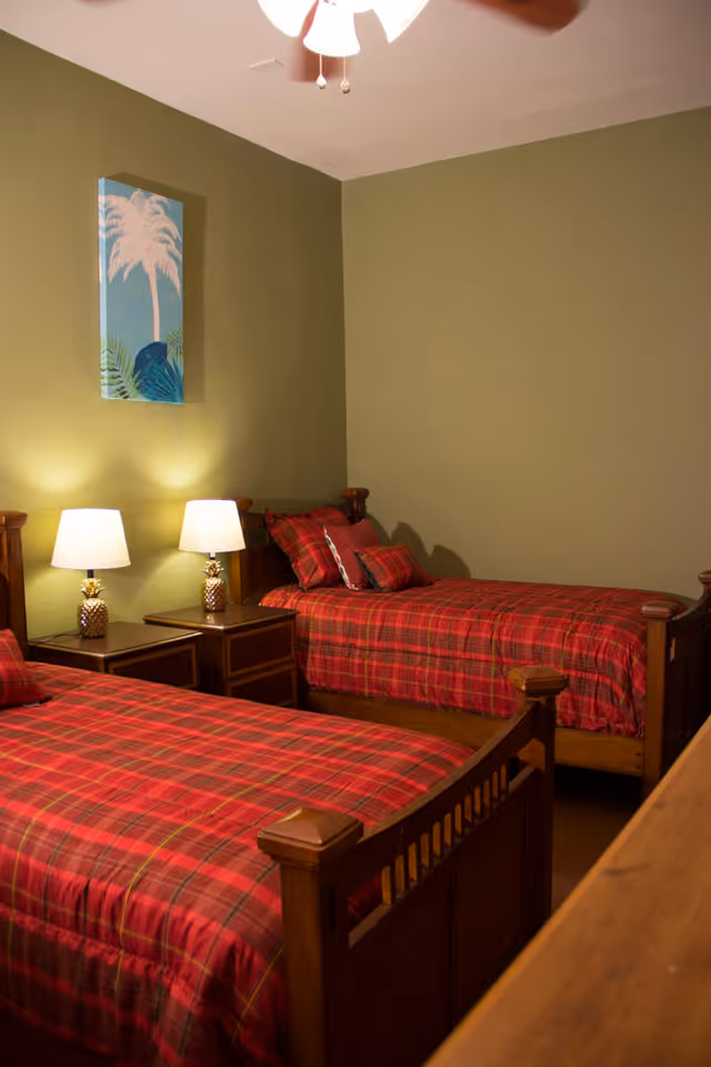A bedroom with two single wooden beds, each covered with red plaid bedding and matching pillows. Between the beds are two nightstands with pineapple-shaped lamps. The walls are painted olive green, and a ceiling fan with lights is visible. A framed artwork featuring a palm tree hangs on the wall above one of the beds.