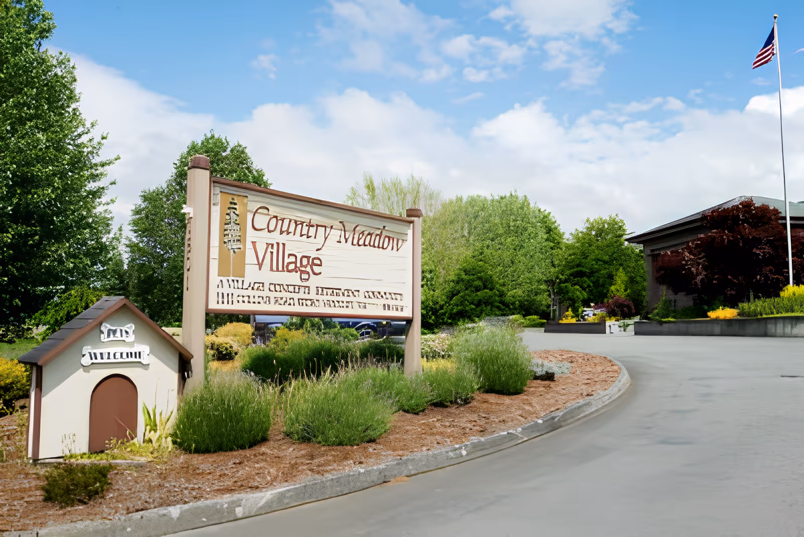 Entrance sign reading 'Country Meadow Village' beside a landscaped driveway and flagpole in front of the facility.