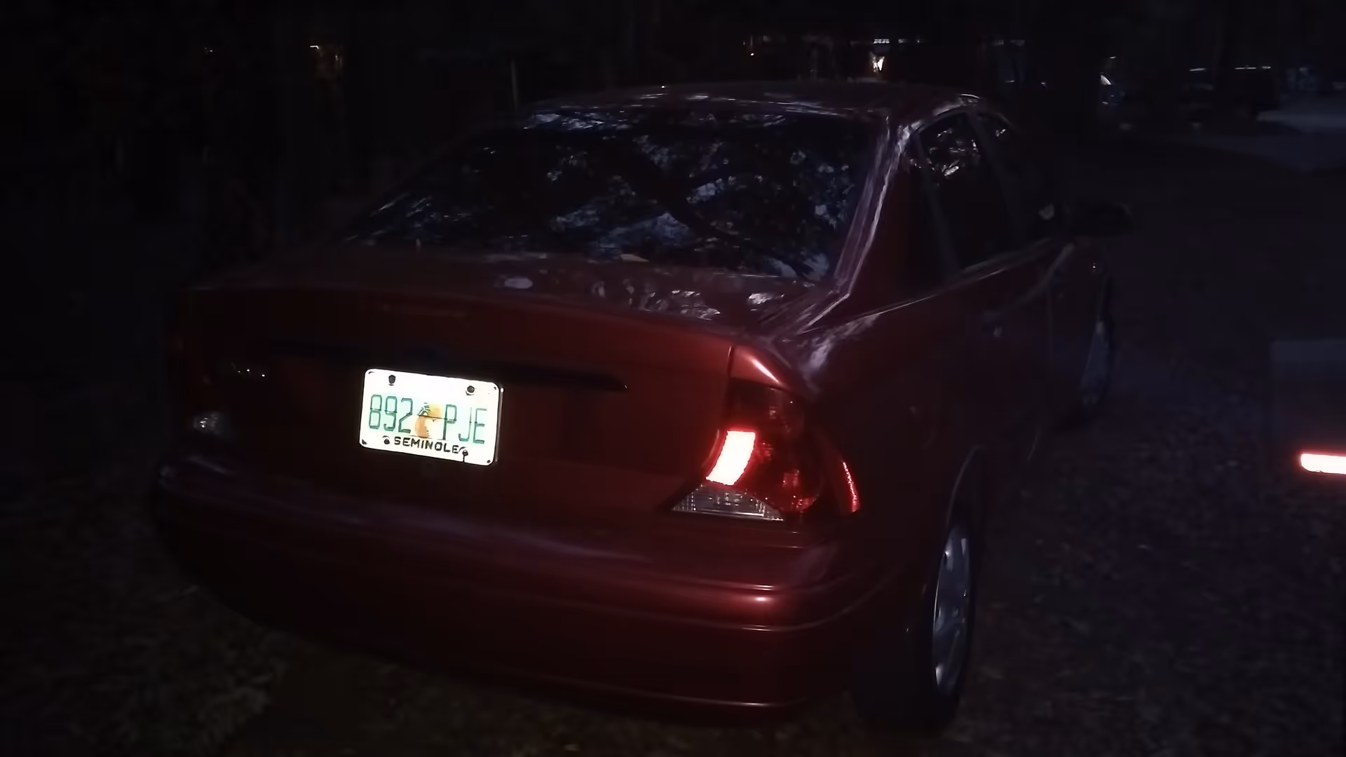 Red sedan parked on a dark gravel lot with illuminated taillights at night.