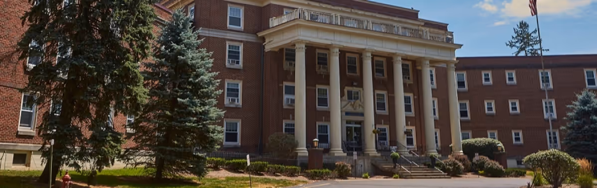 Front exterior of a large brick senior living building with tall white columns, steps at the entrance, surrounding trees, and a flagpole.