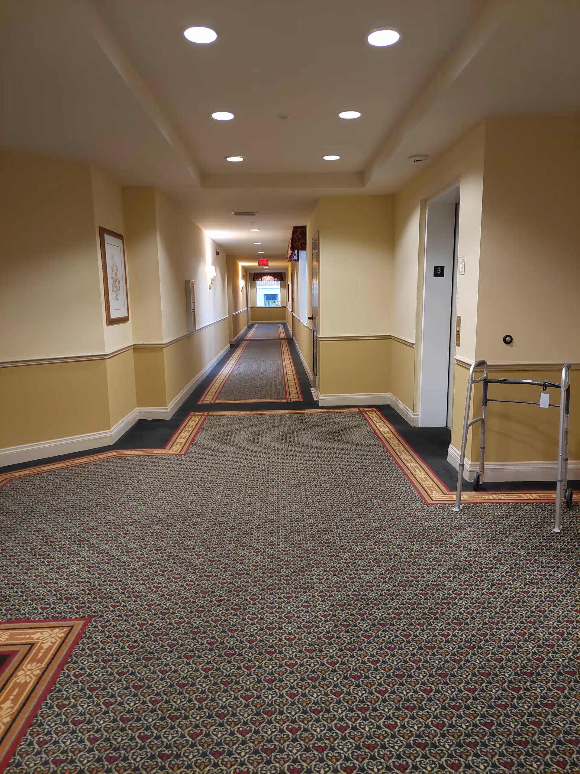 Long beige hallway in a senior living facility with patterned carpet and ceiling lights.