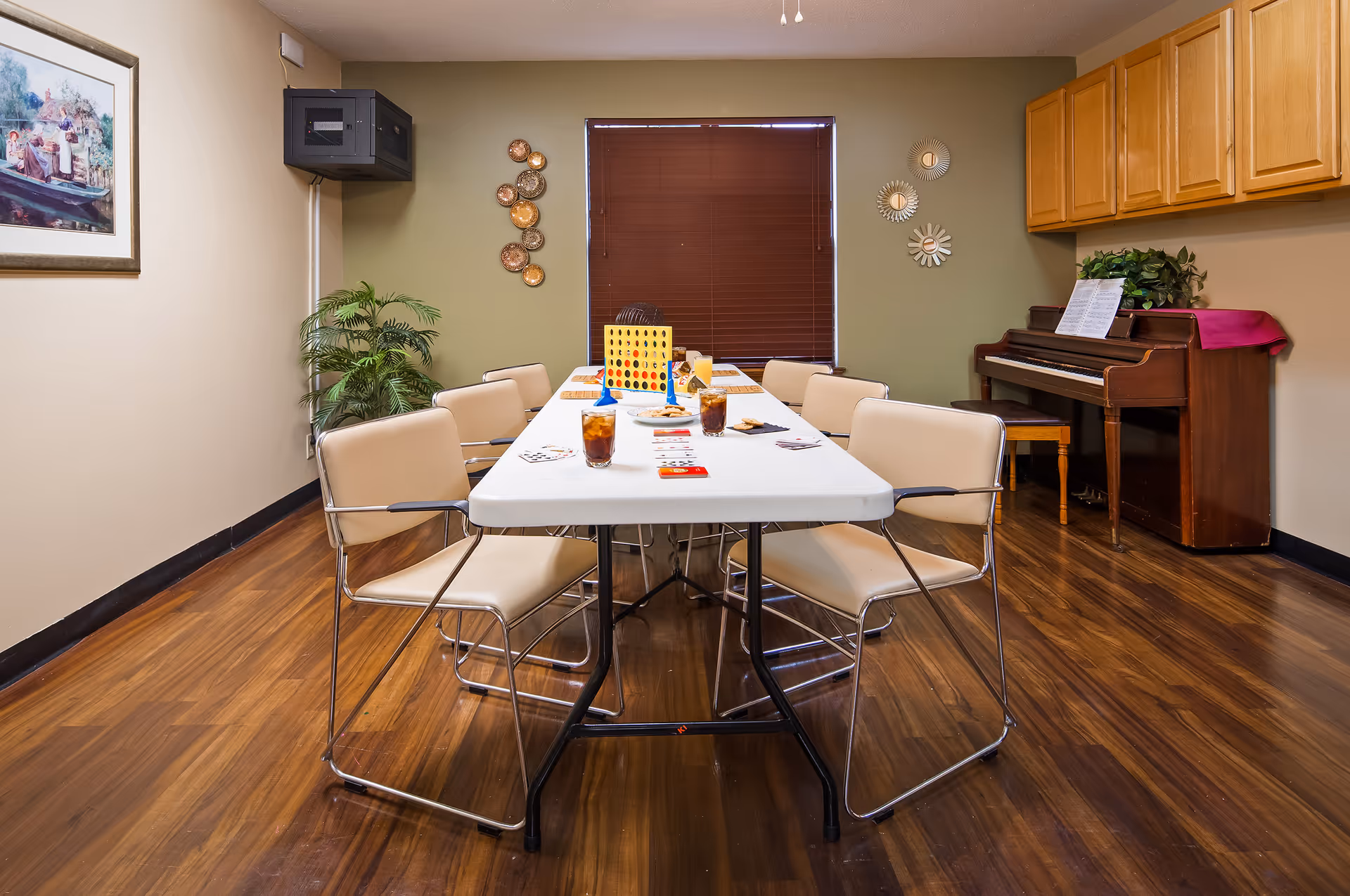 A room with a long white table surrounded by beige chairs on a wooden floor. On the table are playing cards, glasses of iced tea, a plate of cookies, and a Connect Four game. The room has green and beige walls, a window with closed brown blinds, a piano with sheet music and a plant on top, wall decorations, and a framed painting on one wall.