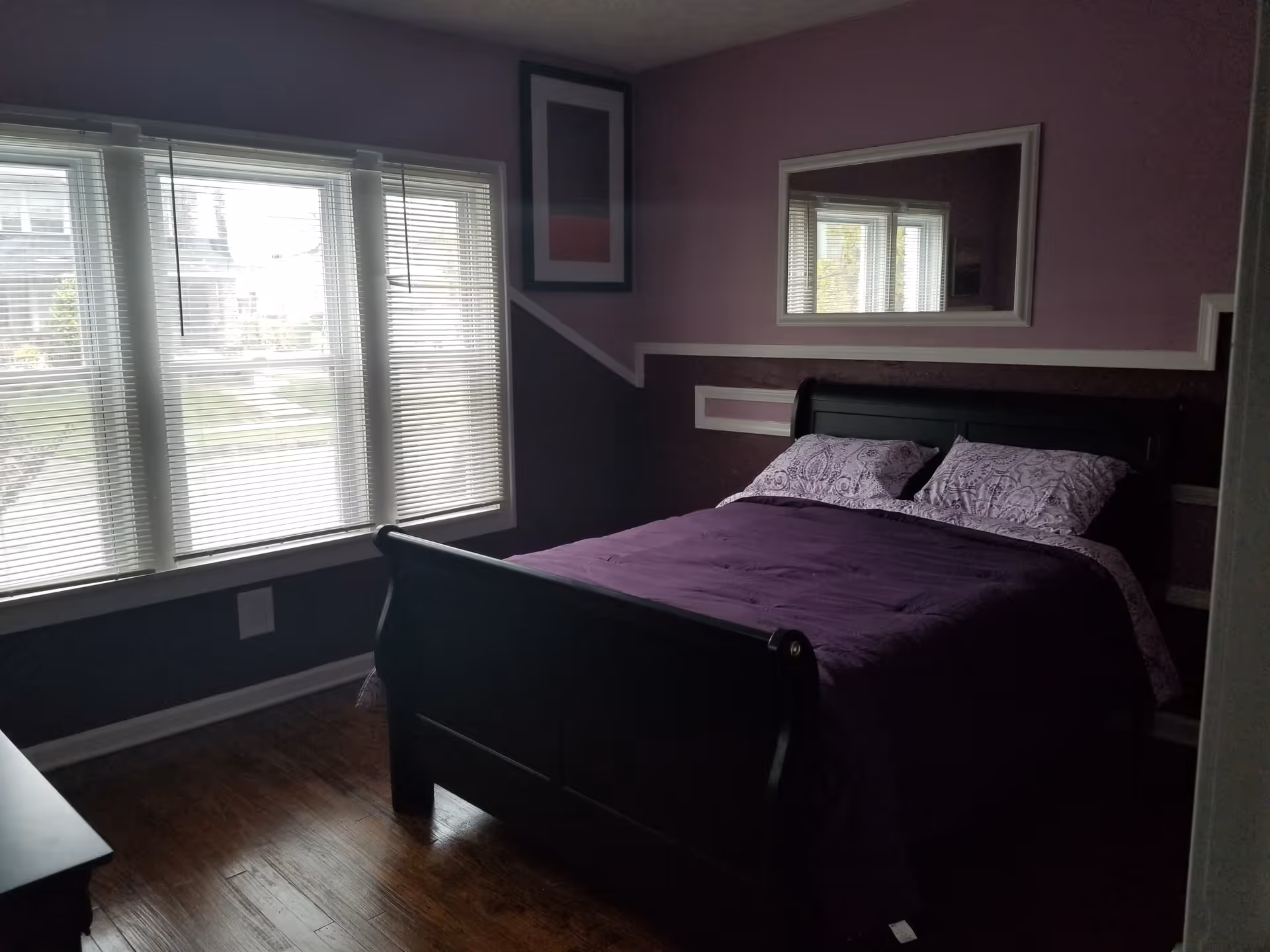 Purple-themed bedroom featuring a bed with purple bedding, large windows with blinds, and a wall mirror.