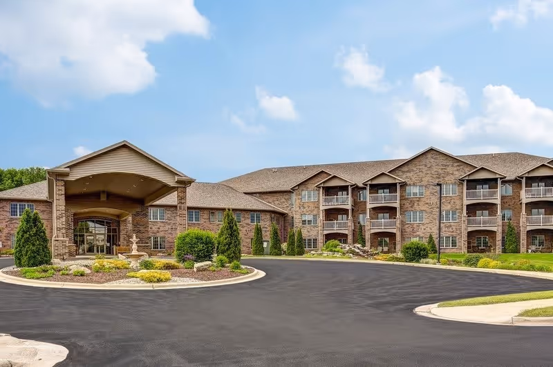 Exterior view of a large brick senior living facility named Brookview Meadows with a covered entrance, landscaped greenery, and a paved driveway under a partly cloudy sky.
