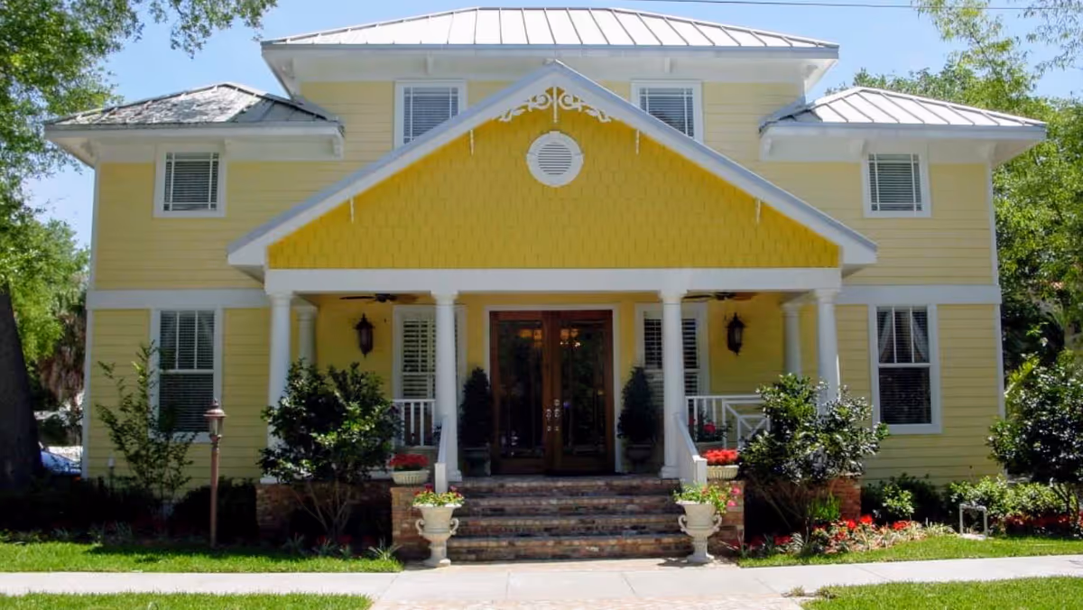 Front exterior view of a two-story yellow building with white trim, a metal roof, and a covered porch supported by white columns. There are steps leading up to double wooden doors, potted plants on either side of the steps, and greenery surrounding the building.