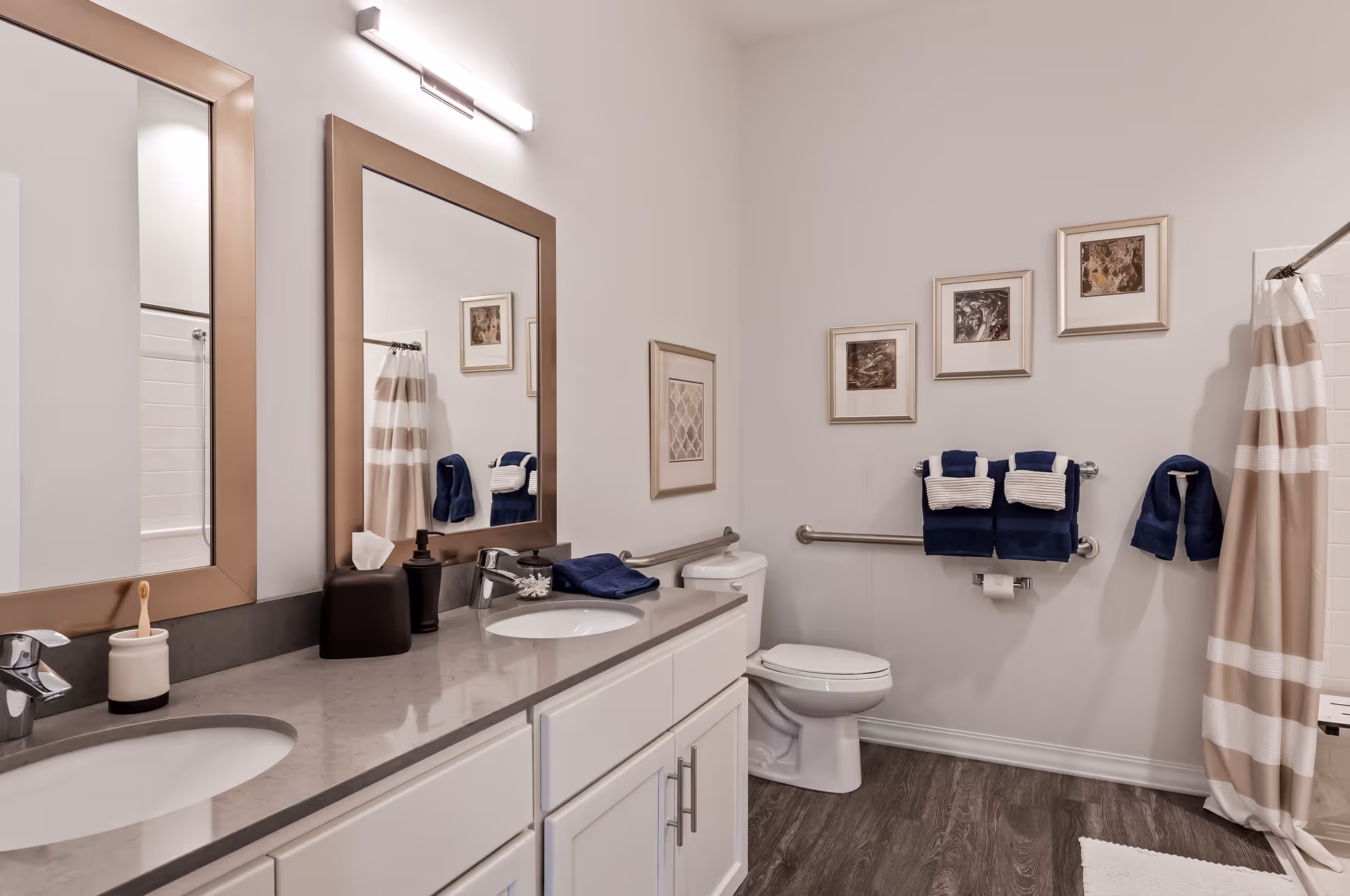A clean and modern bathroom with a double sink vanity featuring two large mirrors and a gray countertop. The bathroom has a white toilet, grab bars on the walls, and a shower with a beige and white striped curtain. Blue and white towels are neatly hung on the towel racks, and framed artwork decorates the walls. The floor is a wood-like finish.