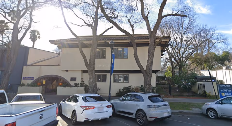 Exterior view of a senior living facility with trees, parked cars, and a gated entrance.