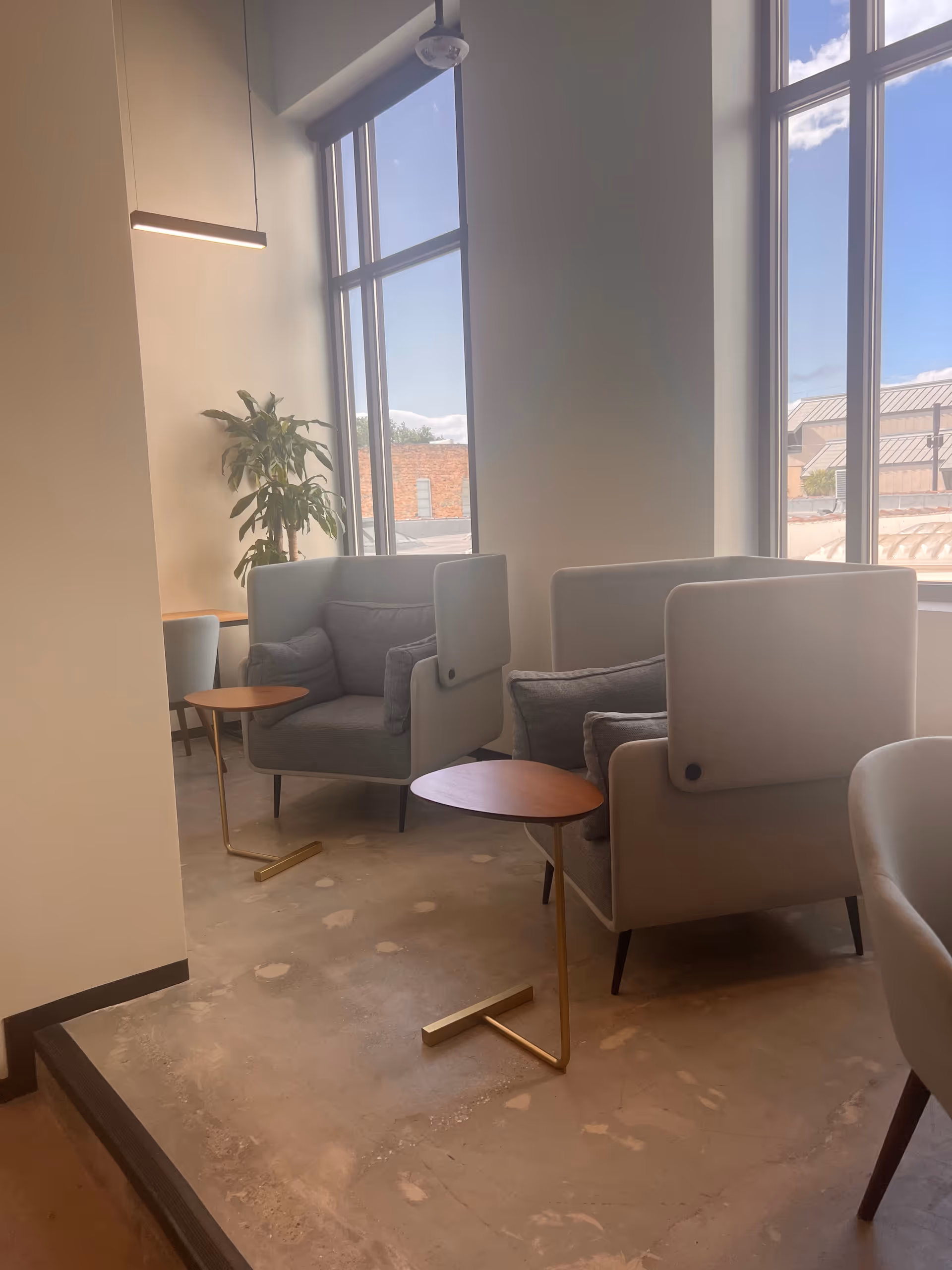 Modern lounge area with gray armchairs, small wooden tables, plant, and large windows.