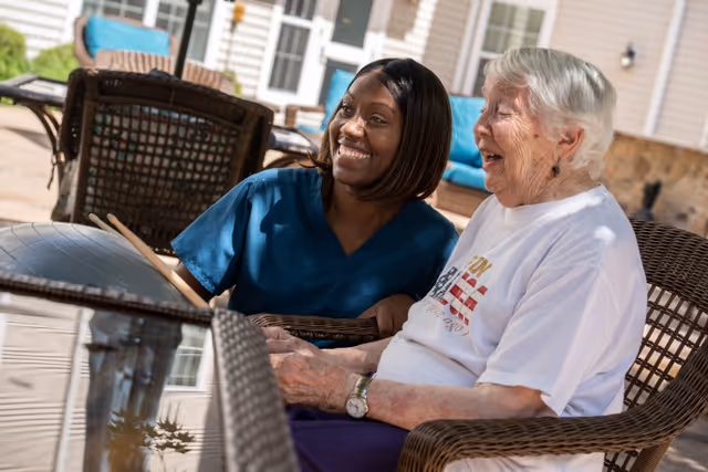 Two women outdoors at a patio, smiling; caregiver with resident near wicker chairs.