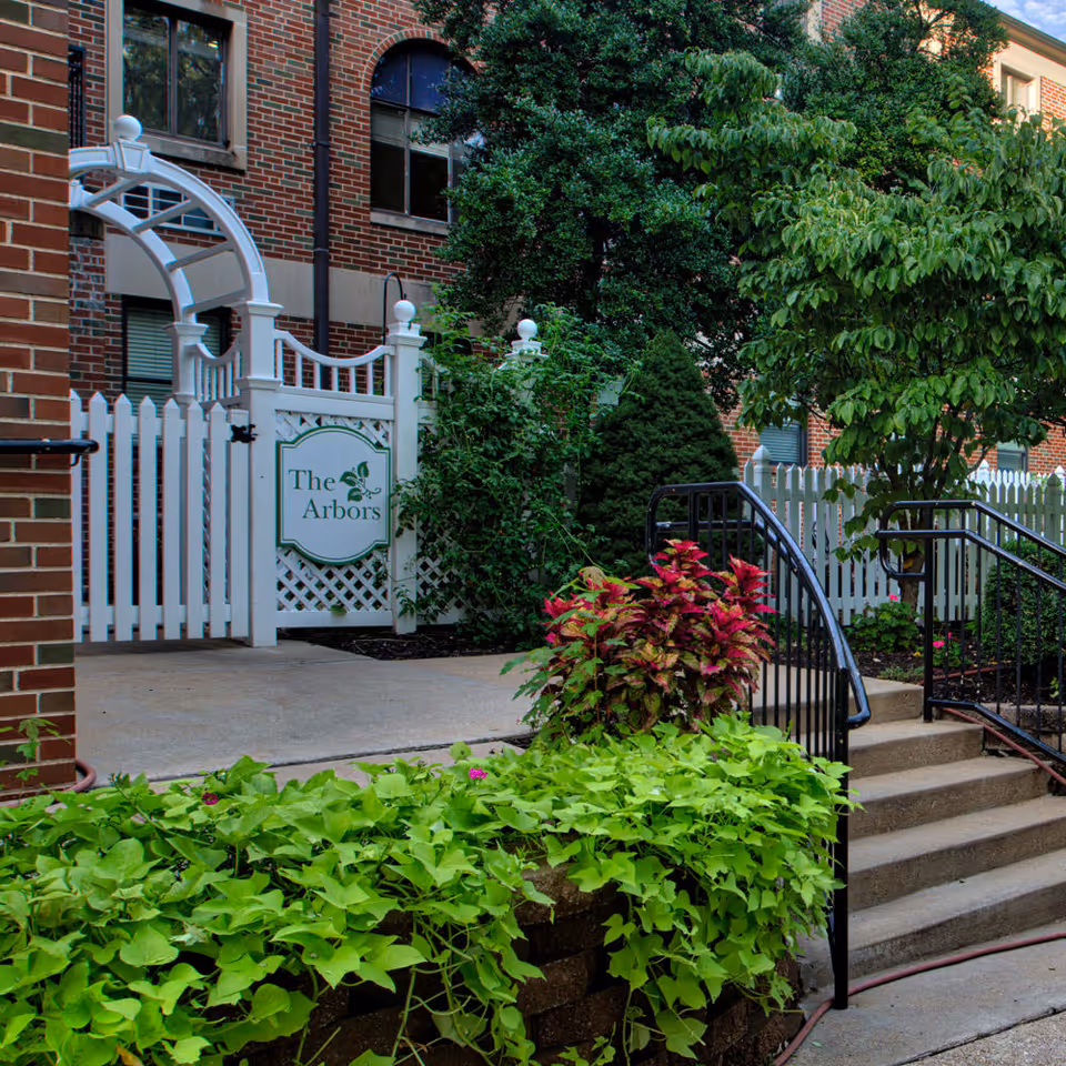 Entryway to 'The Arbors' with a white picket gate and sign, brick building facade, steps, and landscaped greenery.