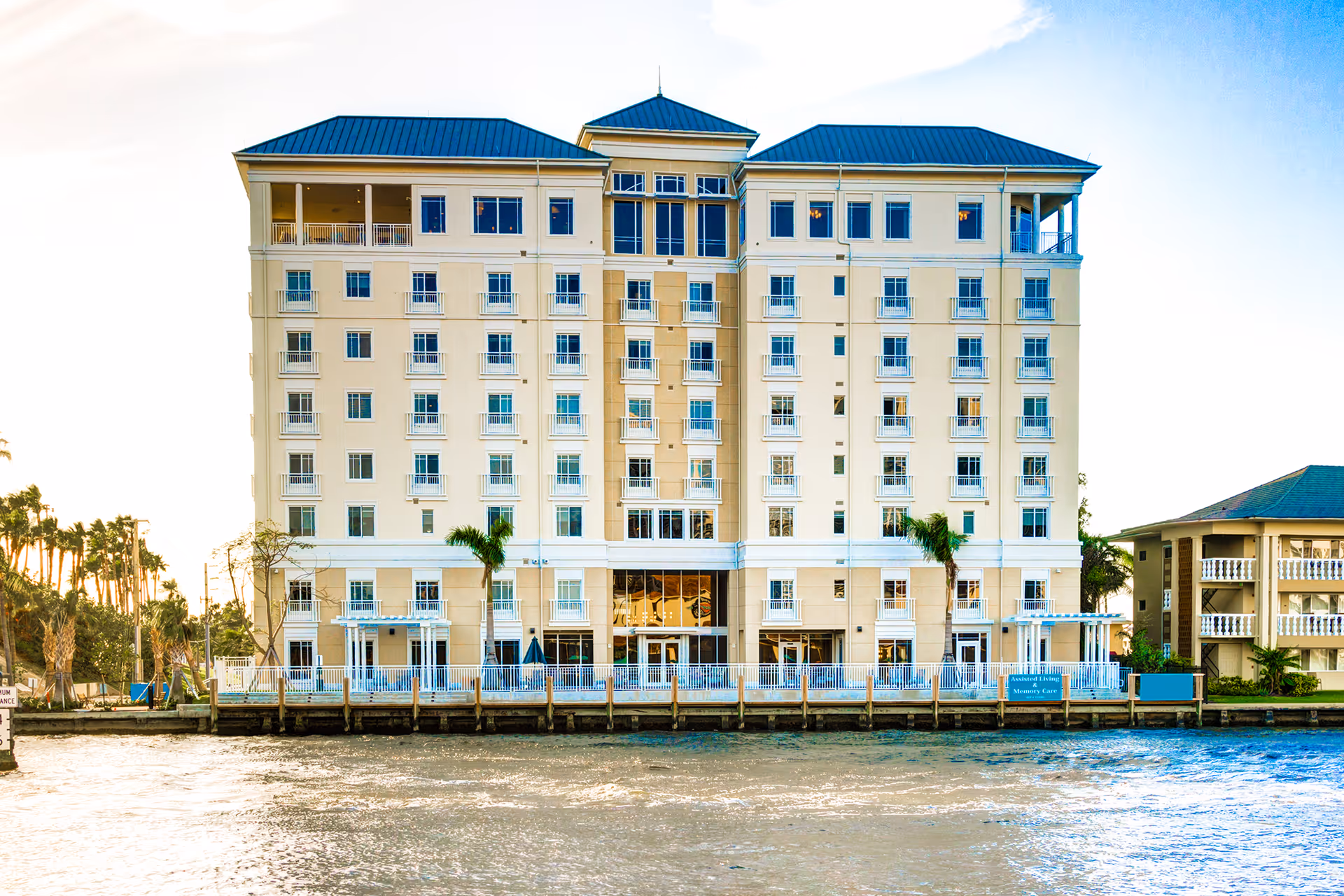 Exterior view of a beige multi-story senior living building by a waterway with balconies